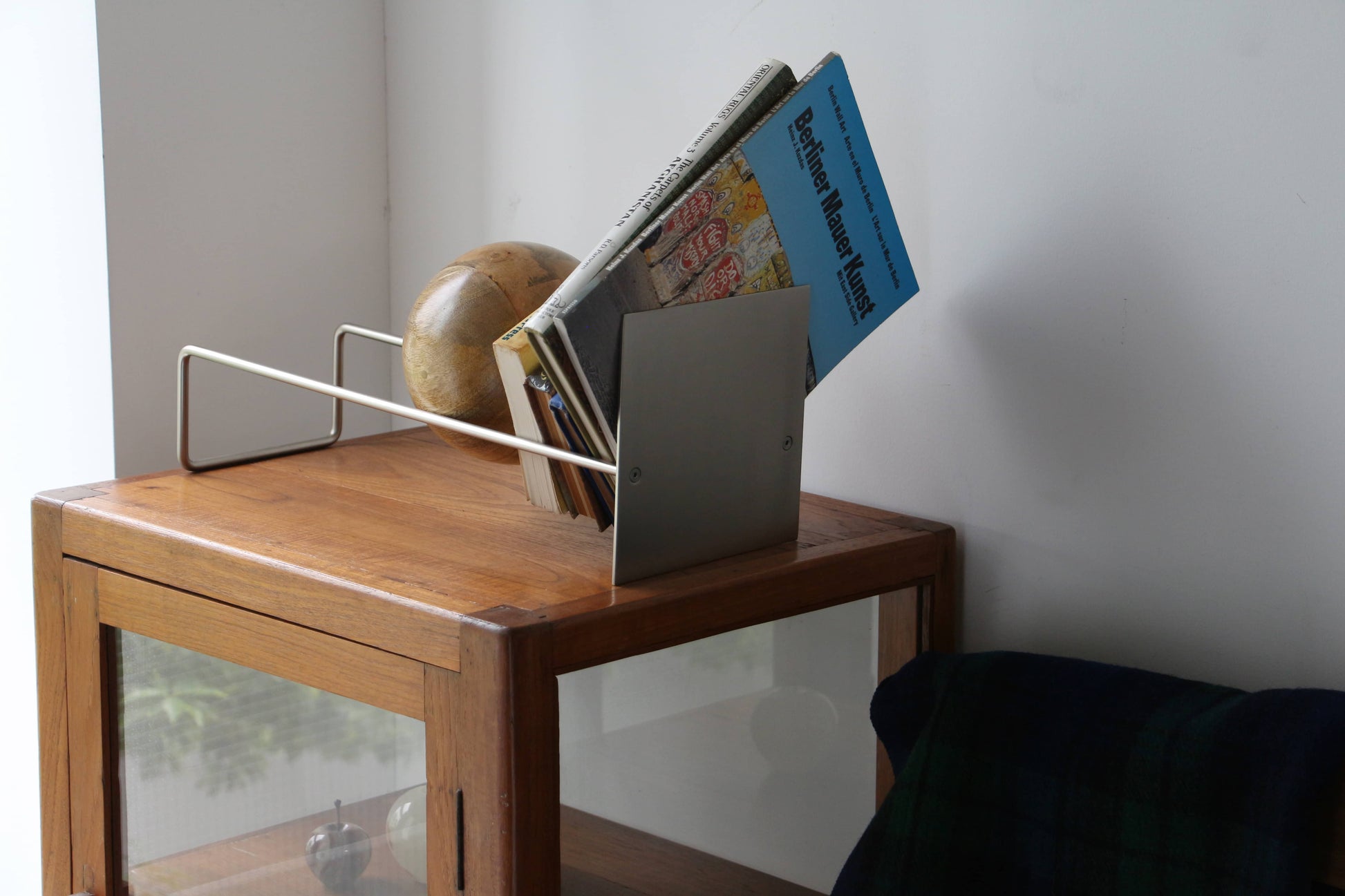 The DETAIL Slope Bookend by DETAIL, a sleek silver steel piece for organizing books and magazines, sits atop a wooden cabinet beside a globe and a dark blanket draped over a nearby chair.