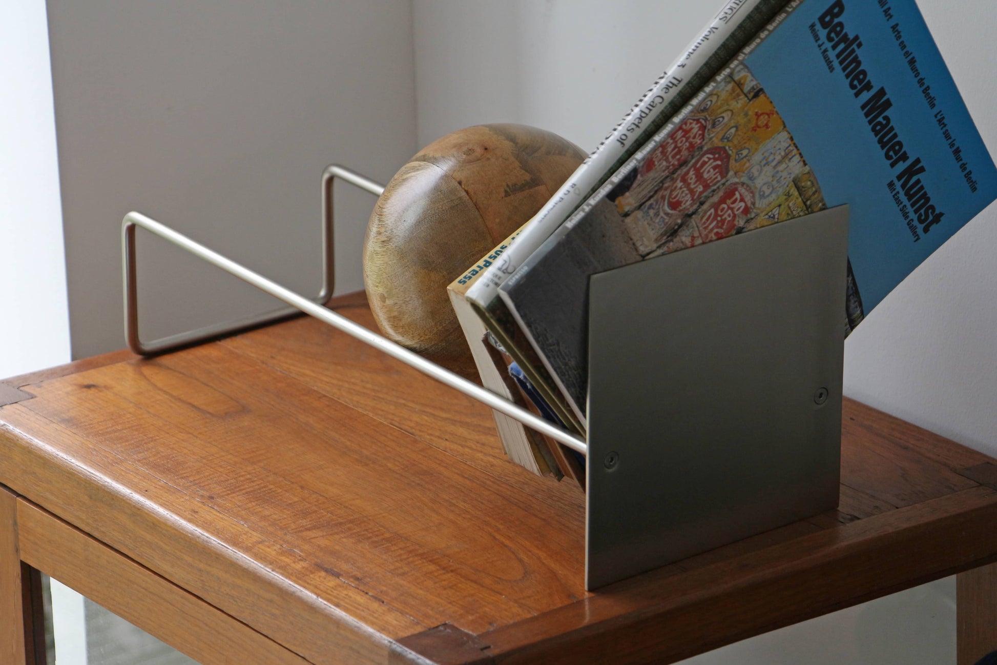 The DETAIL Slope Bookend by DETAIL, featuring a sleek silver steel frame, organizes books and magazines on a wooden table by the window, with a unique mango wood ball globe placed behind the reading materials.