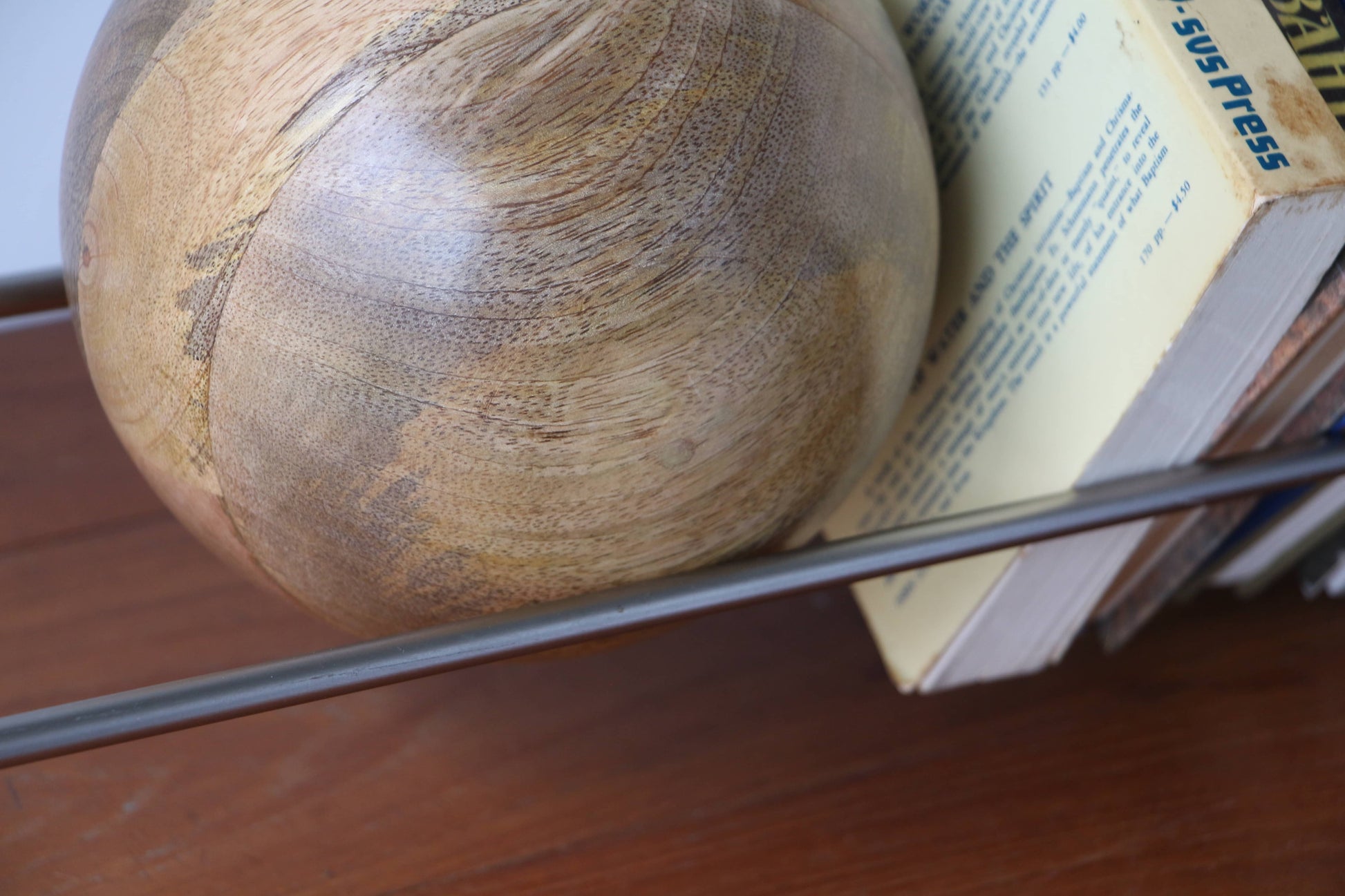 A unique mango wood ball sits beside books held upright by the DETAIL Slope Bookend from DETAIL, crafted in sleek silver steel on a wooden surface. One yellow book with blue text on its spine is visible.