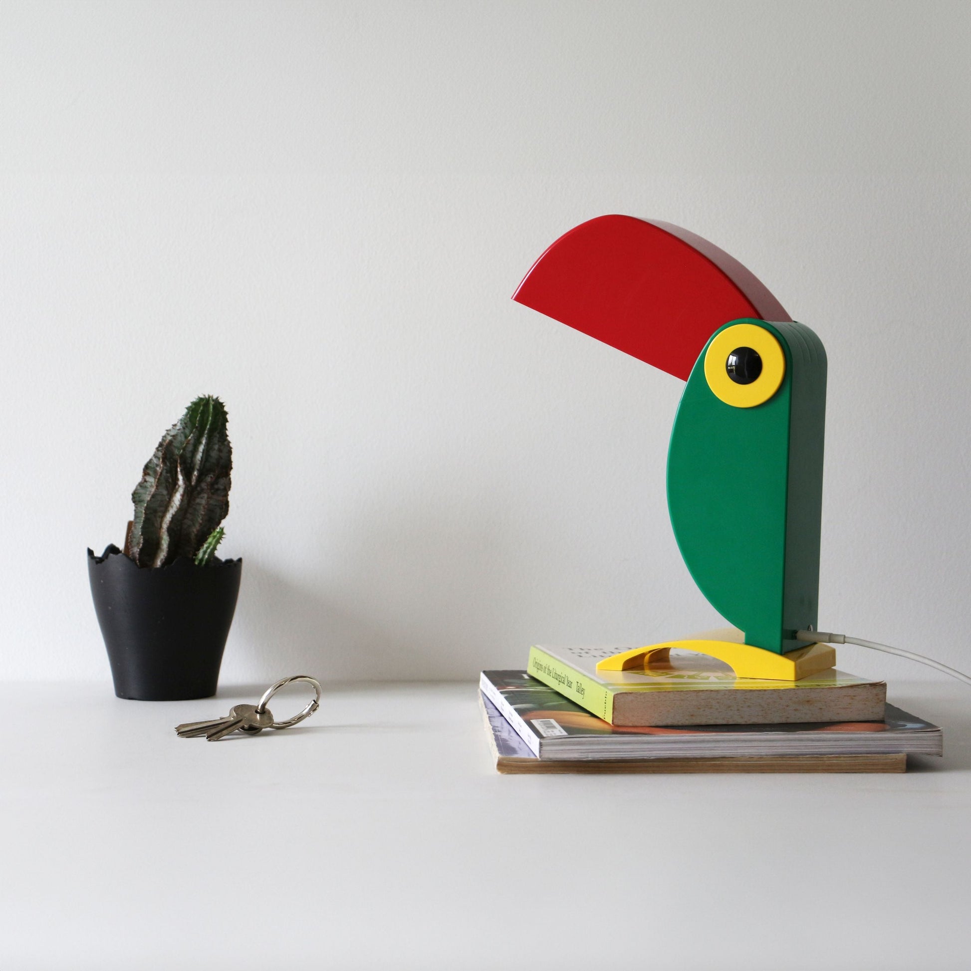 The DETAIL Toucan Lamp by DETAIL, featuring a red beak and green body with iconic vintage charm, sits on a stack of books. A small potted cactus and keys are nearby on a white surface against a plain background.