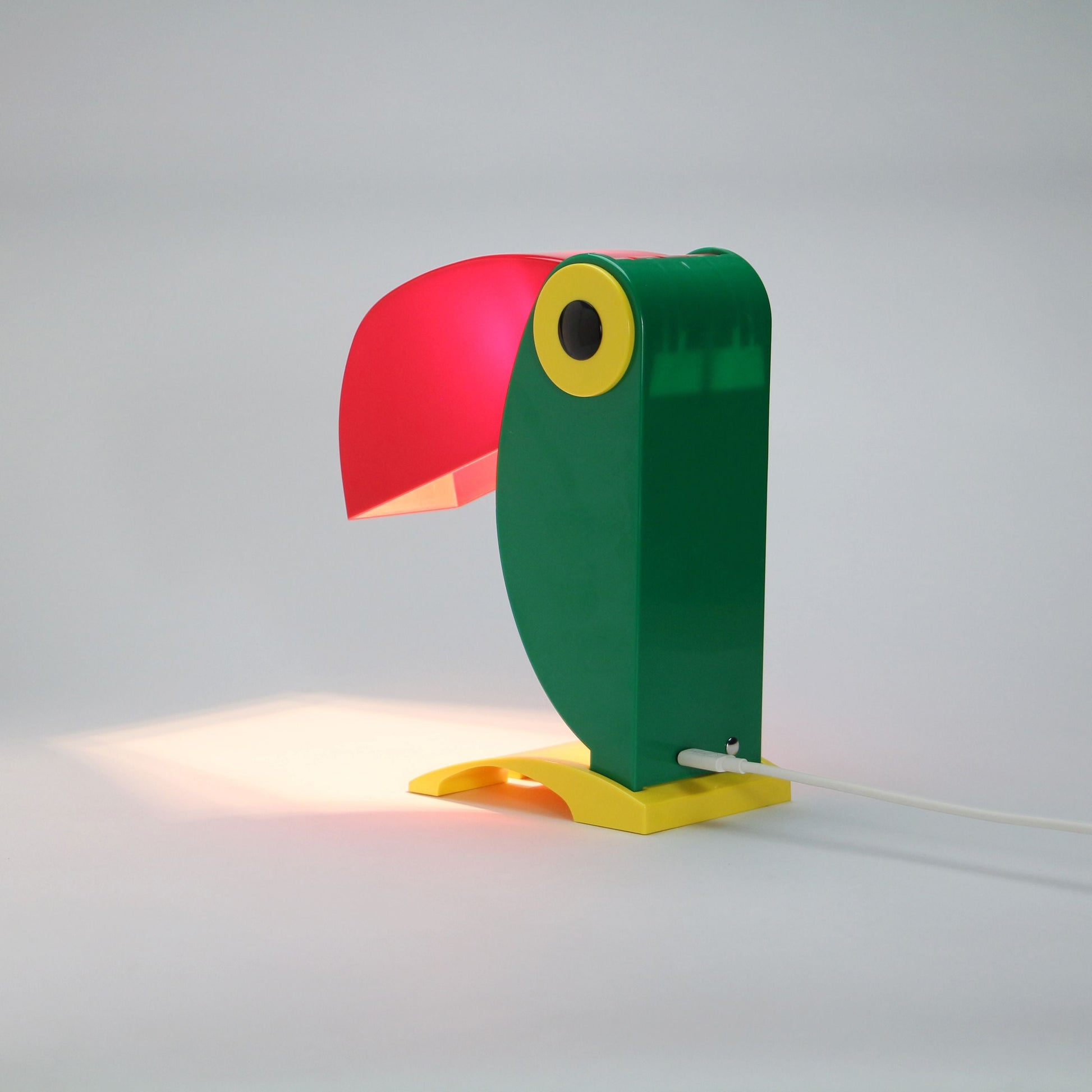 The DETAIL Toucan Lamp by DETAIL features a green body, yellow base and eye, and a red beak lampshade—an iconic vintage design that casts a warm glow onto any white surface.