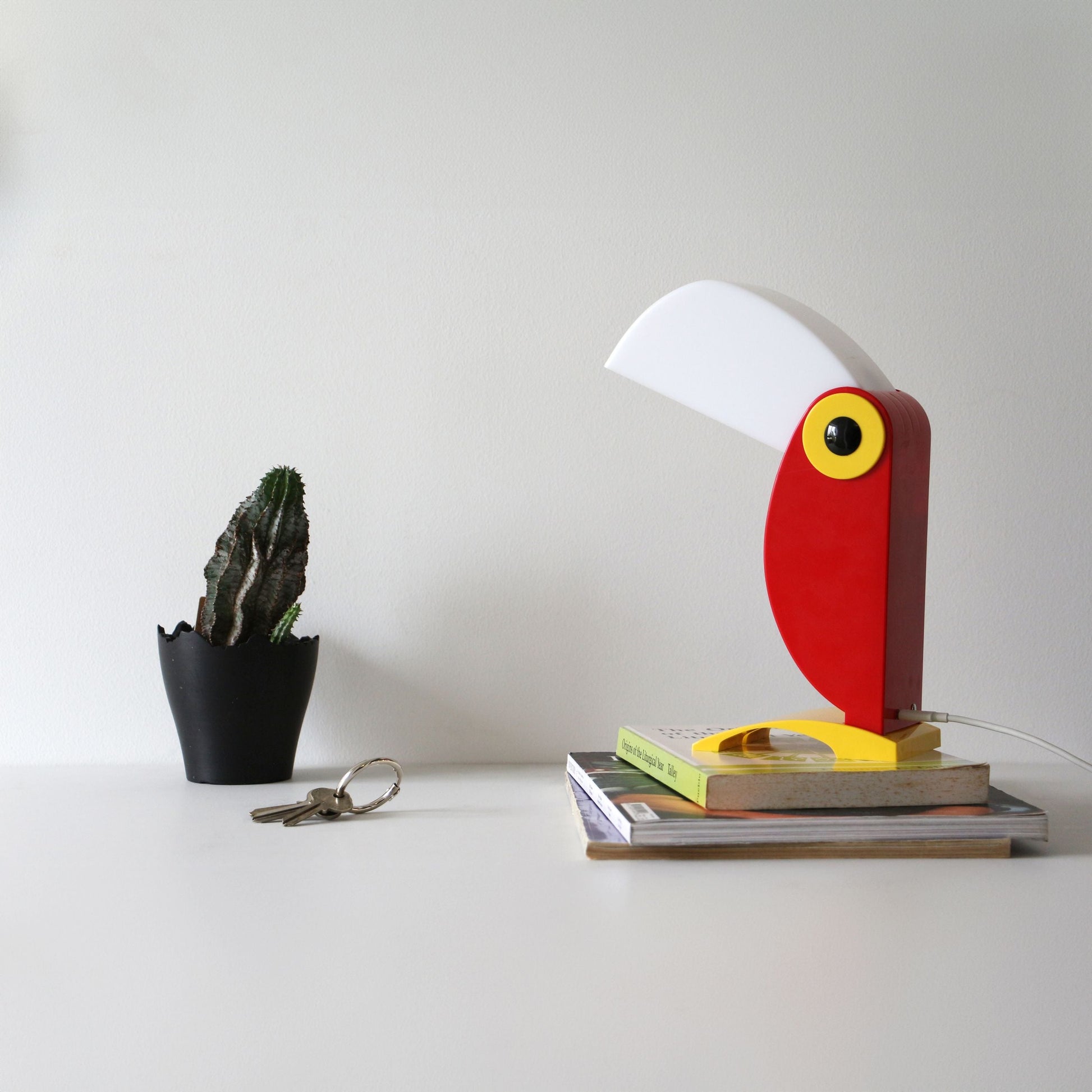 A desk with a small potted cactus, a set of keys, and the DETAIL Toucan Lamp by DETAIL featuring its iconic design, arranged on stacked books against a plain white wall.