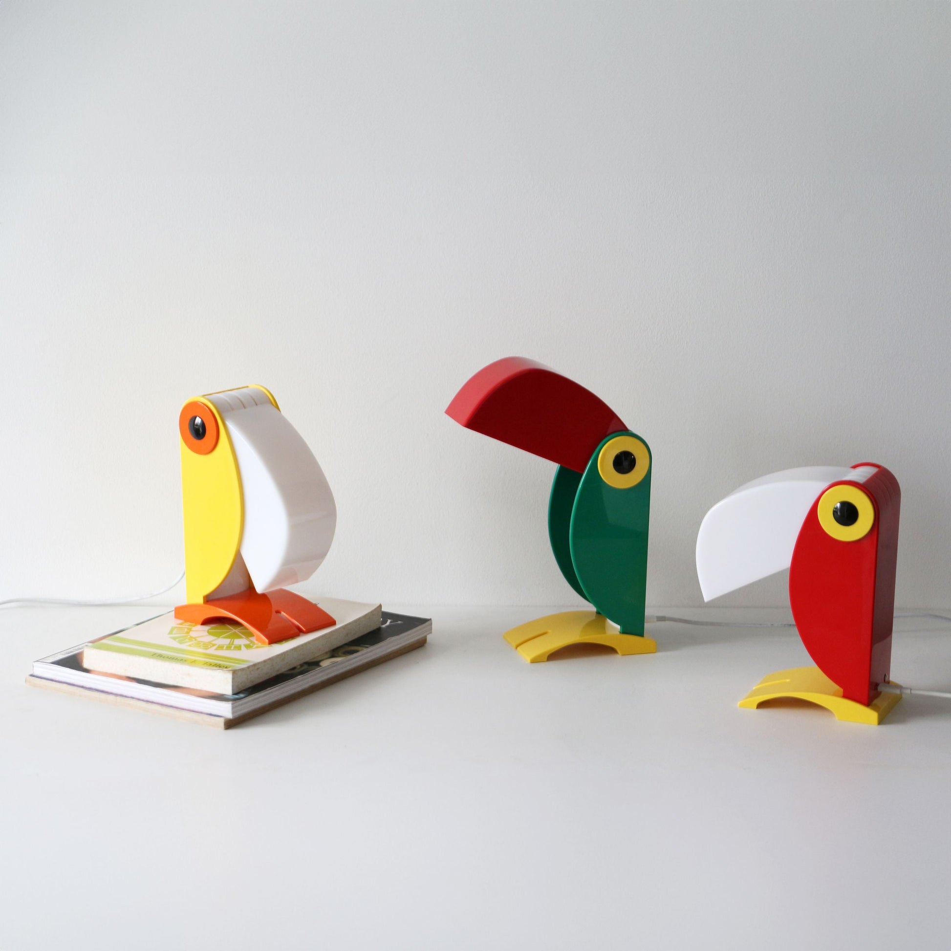 Three colorful DETAIL Toucan Lamps by DETAIL stand on a white surface, each exuding vintage charm—one yellow and white resting on a closed book, one green and red, and one red and white—highlighting their iconic beak-shaped lamp covers.