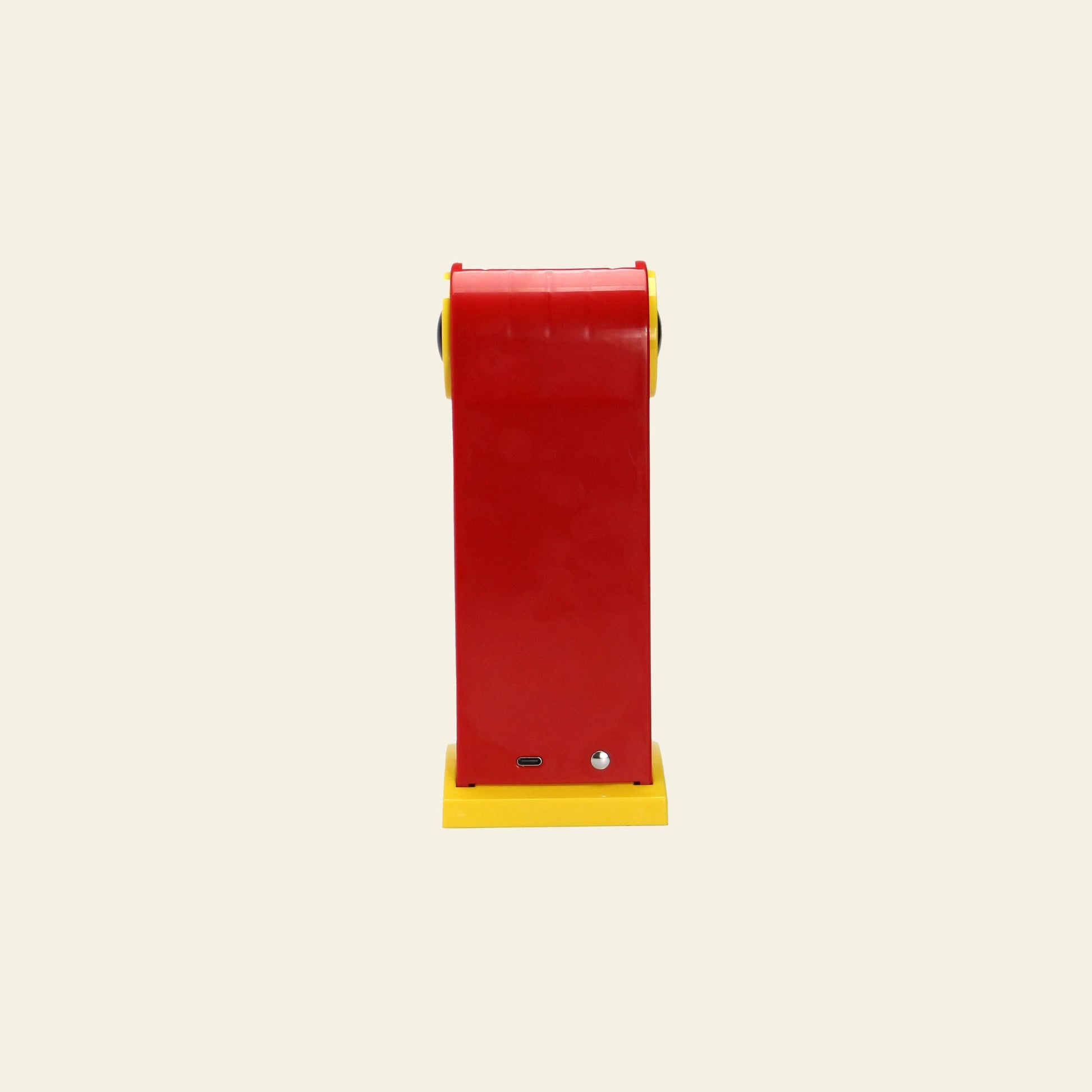A DETAIL Toucan Lamp by DETAIL stands upright against a plain beige background, featuring a red rectangular body with yellow edges and base, a small front port, and a silver button—echoing its iconic classic design.