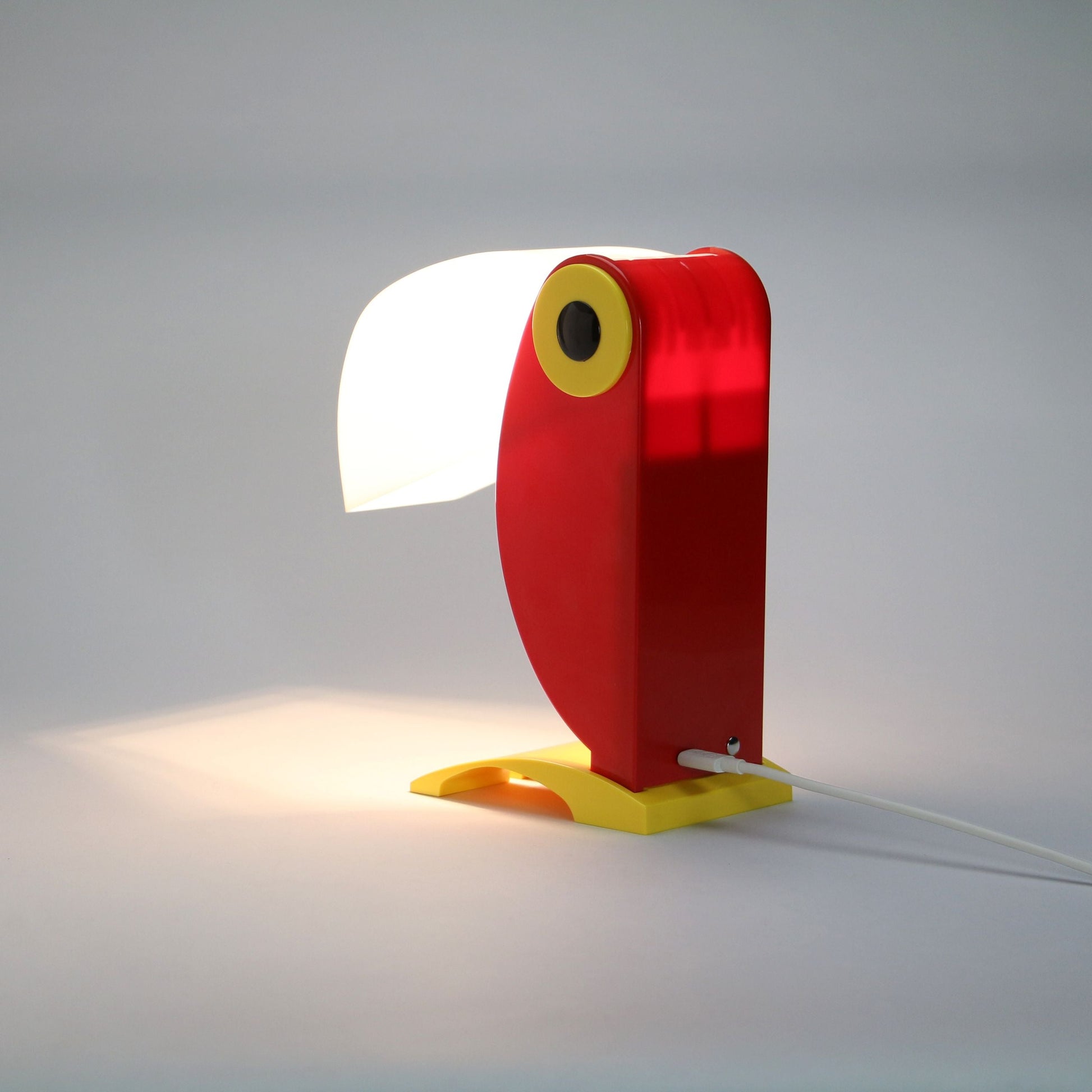 The DETAIL Toucan Lamp by DETAIL adds vintage charm with its red body, yellow beak-inspired base, and distinctive white beak-shaped lampshade that casts light to the side in an iconic design.