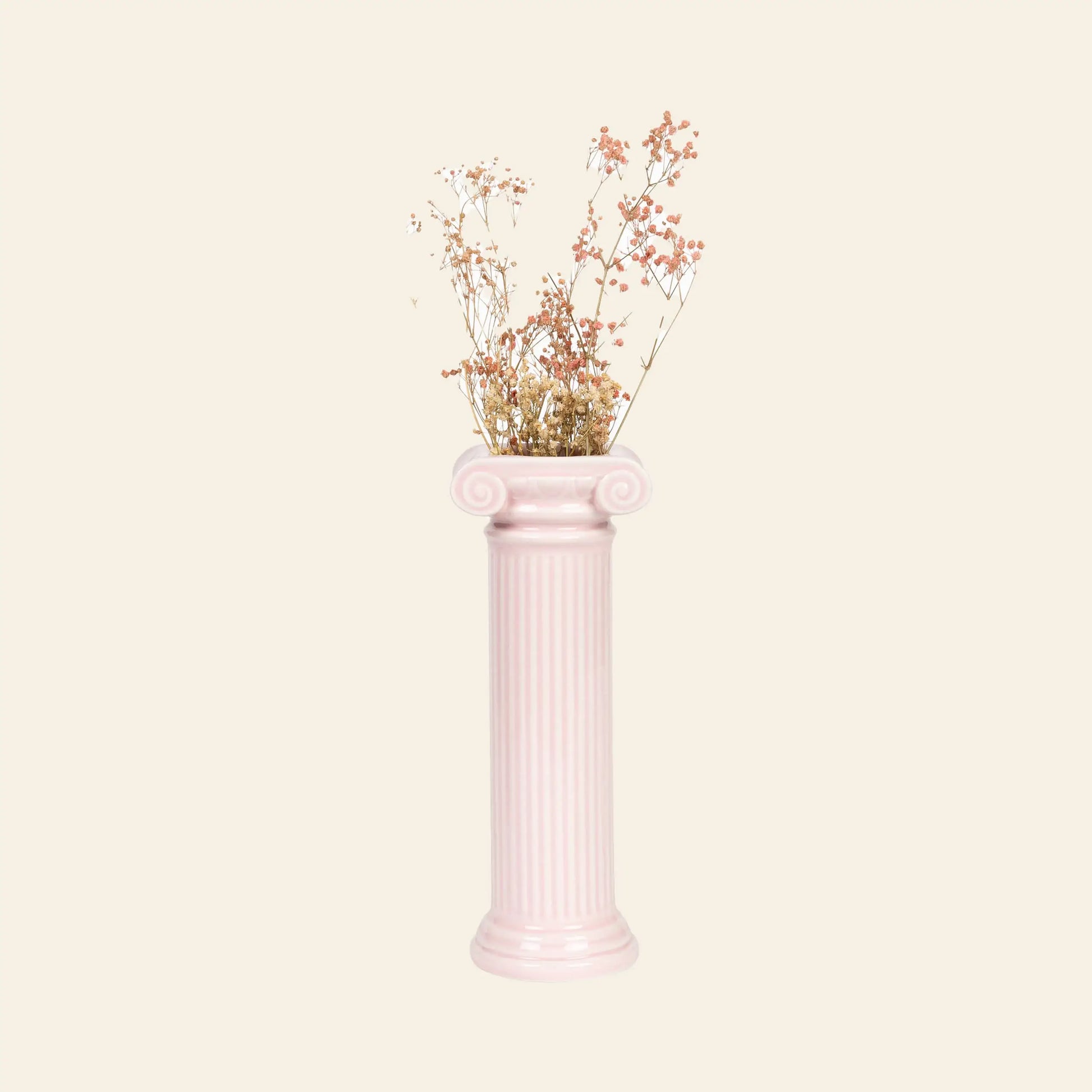 The DOIY Athena Vase by DOIY, a tall pale pink ceramic piece inspired by a Greek Ionic column, displays delicate dried flowers with small pink and beige blossoms against a plain beige background.