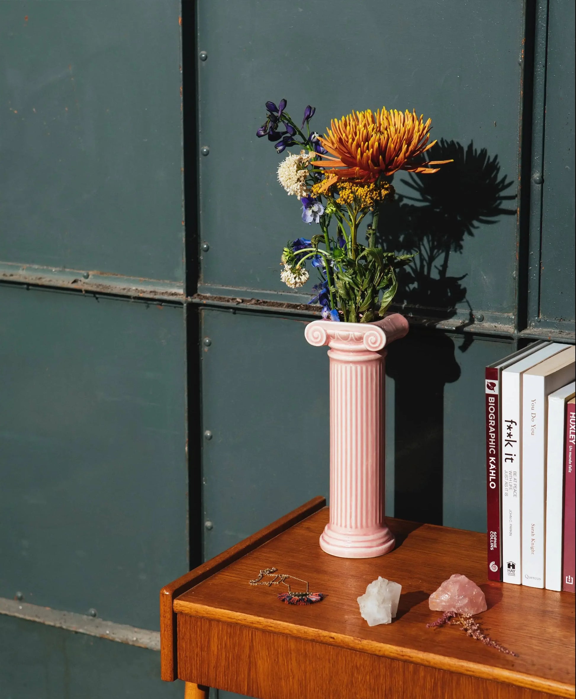 The DOIY Athena Vase by DOIY, inspired by a Greek Ionic column, displays colorful flowers on a wooden table with books, jewelry, and crystals. Sunlight casts shadows on the dark green industrial-style wall.