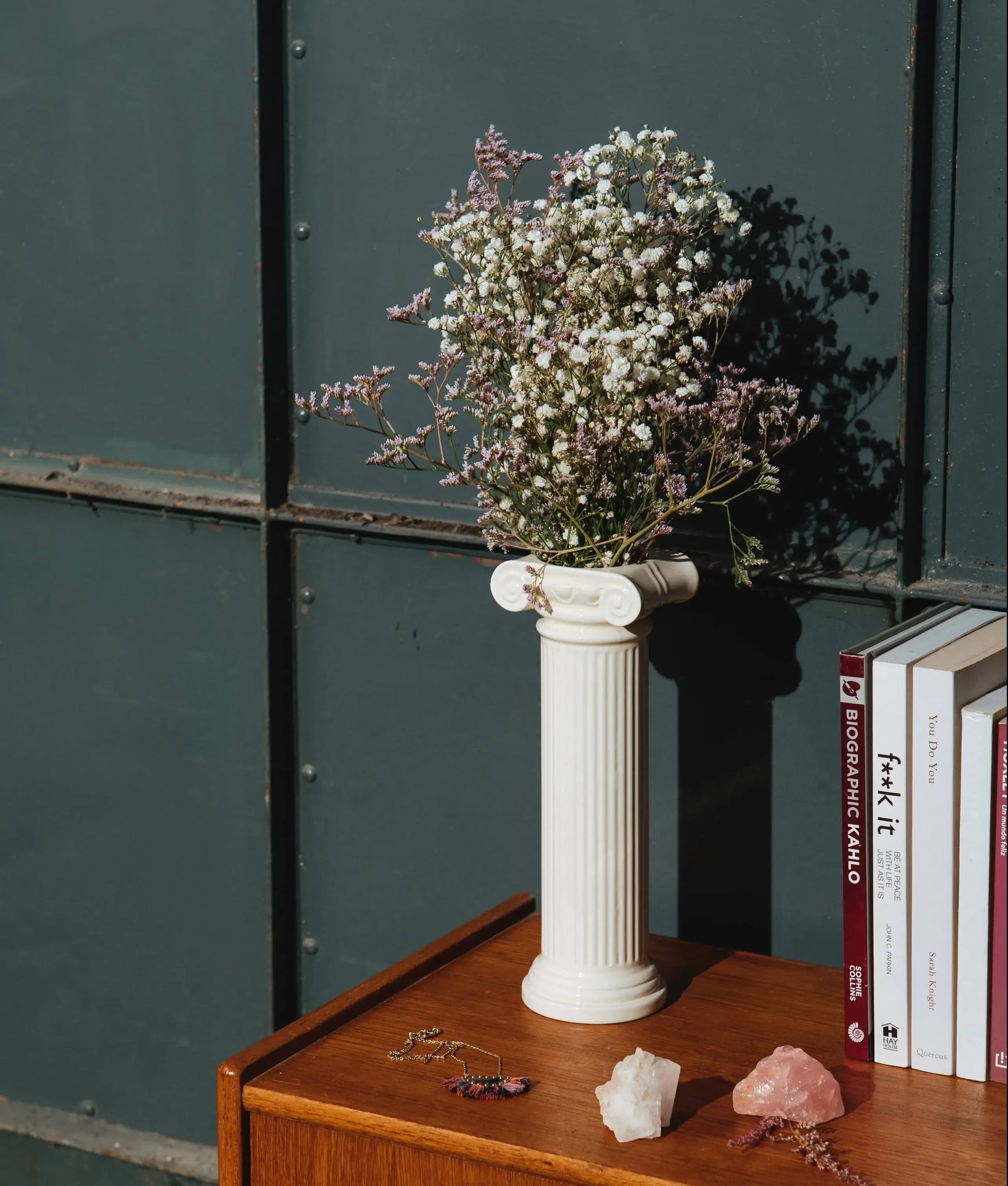 The DOIY Athena Vase by DOIY, styled as a Greek Ionic column in white, holds white and pink flowers on a wooden table with books, crystals, and jewelry nearby against a dark paneled backdrop.