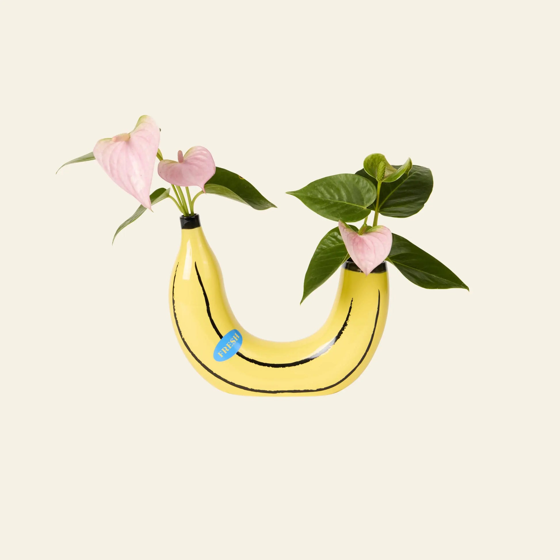 The DOIY Banana Vase by DOIY features a curved design with a blue sticker, holding green leaves and pink anthuriums for a playful home decor touch, set against a plain beige background.