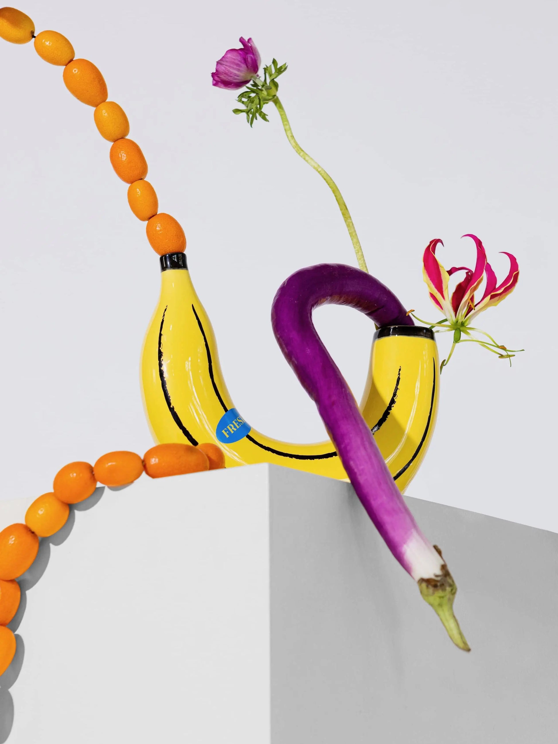 The DOIY Banana Vase by DOIY, styled as a dual vessel, displays a purple eggplant and vibrant flowers. Orange kumquats add a playful touch to this home decor piece, set against a clean white and gray backdrop.
