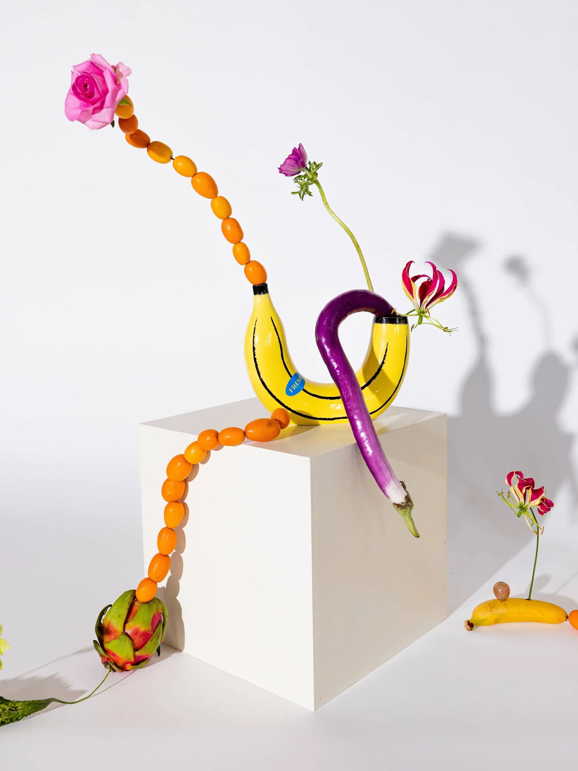 A surreal home decor scene: the DOIY Banana Vase by DOIY, bananas, orange kumquats, pink roses, and other flowers are artistically arranged on a white cube against a white background.