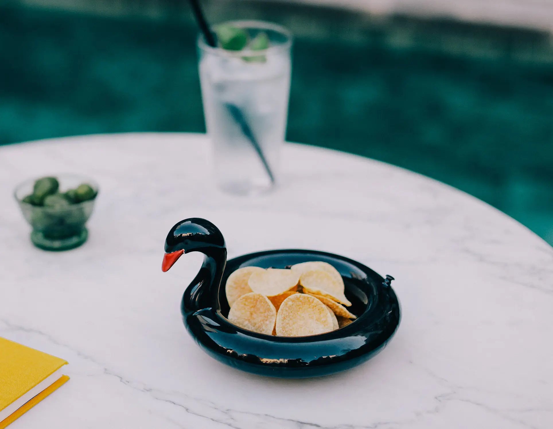 The DOIY Black Swan Floatie Bowl by DOIY, filled with potato chips, rests on a round white marble table near the pool next to a glass of water with a straw and a small dish of green olives.