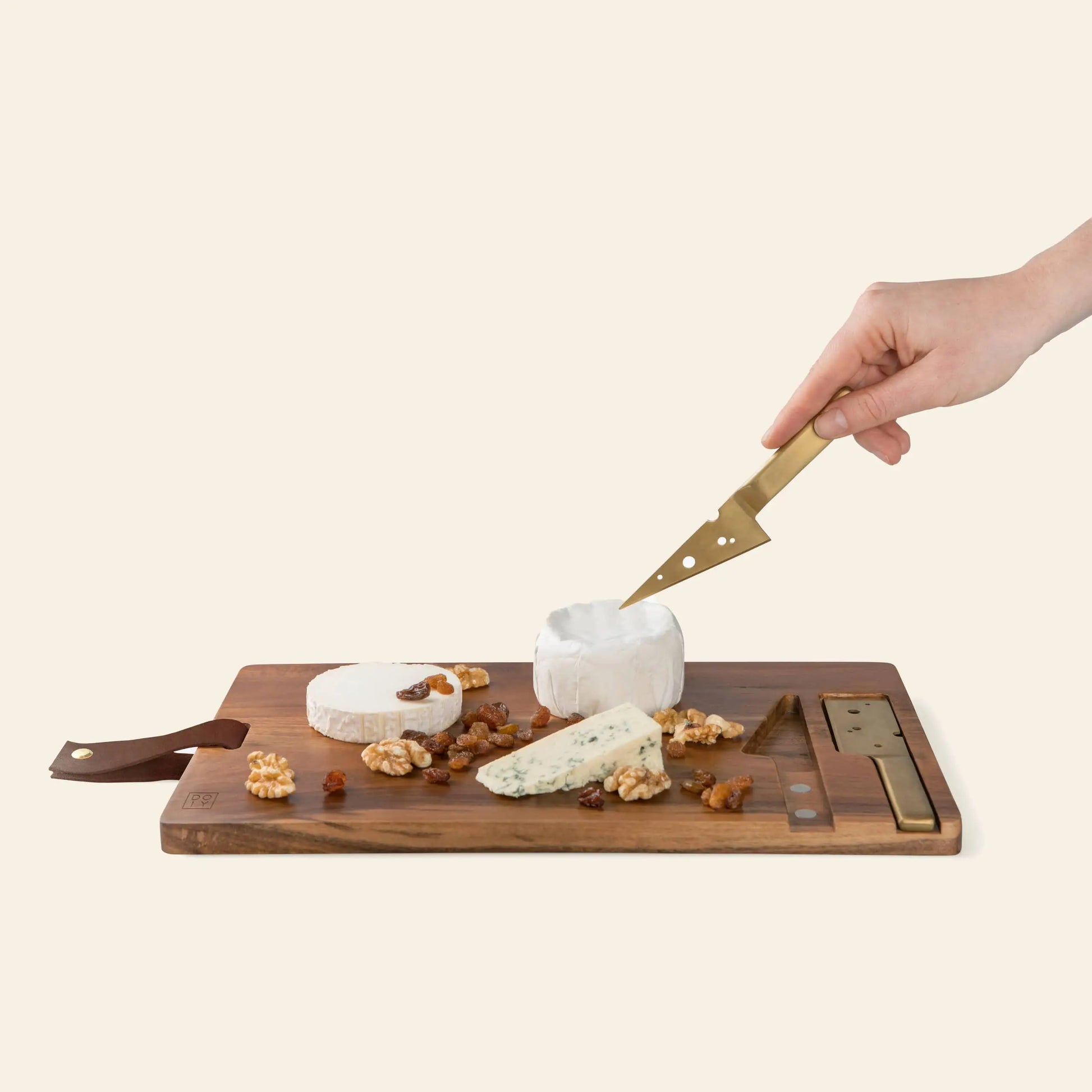 A hand uses the DOIY Cheeseporn cheese knife to slice a round of soft cheese on an Acacia wood board by DOIY, served with two additional cheeses, walnuts, and dried fruit.