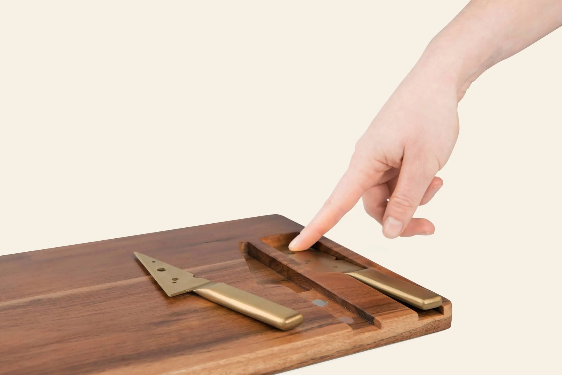 A hand points toward the DOIY Cheeseporn by DOIY, an Acacia wood cheeseboard featuring two golden knives resting in grooves, set against a plain, light background.