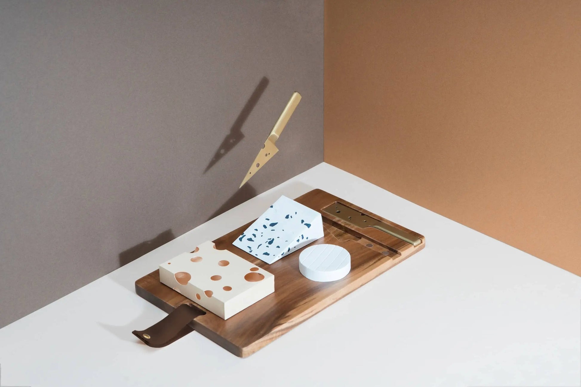 The DOIY Cheeseporn cheeseboard by DOIY, crafted from acacia wood, presents three cheese blocks, a slicer, and a floating golden knife with shadow highlights against a brown and beige backdrop.