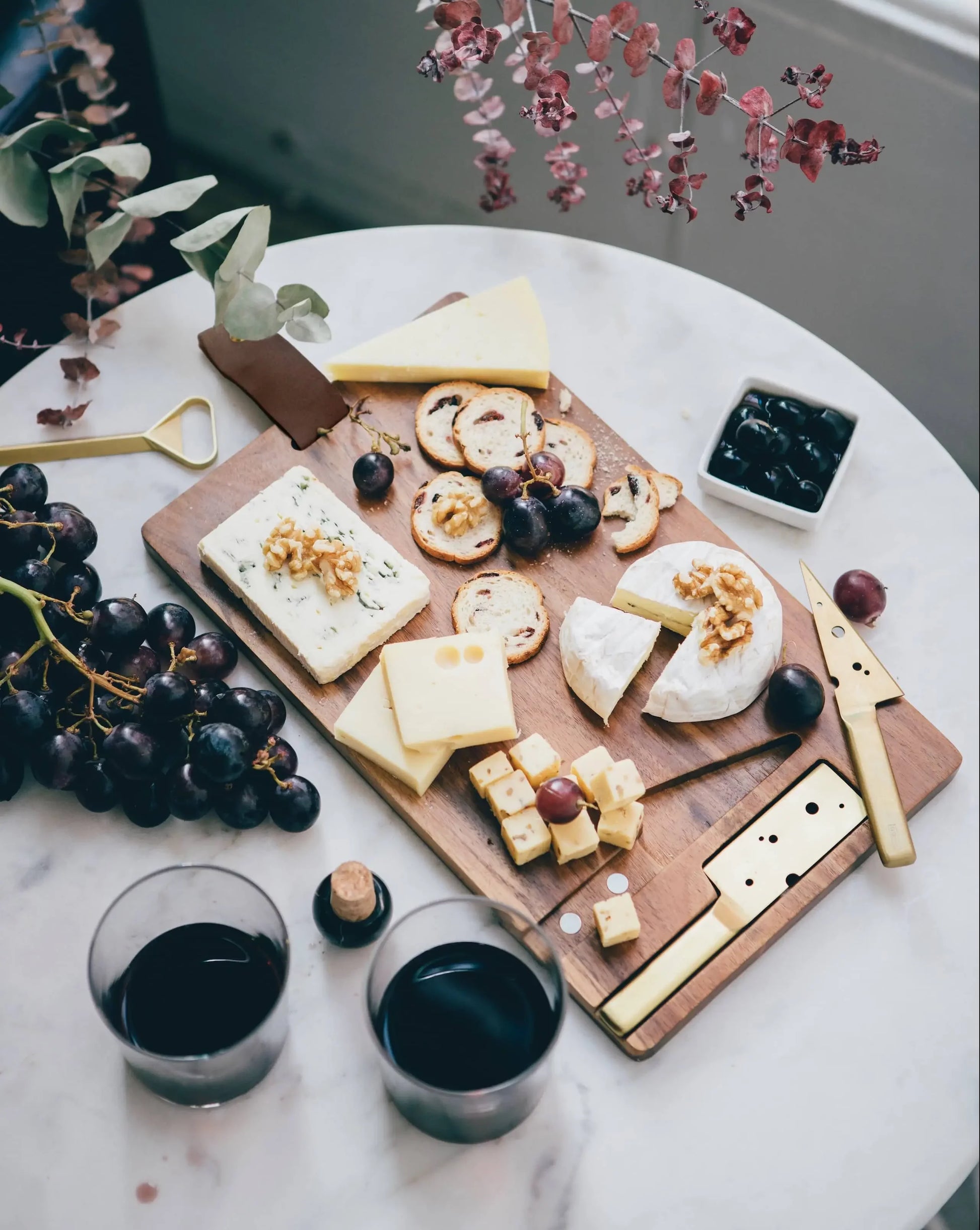 The DOIY Cheeseporn by DOIY features a variety of cheeses, crackers, grapes, nuts, olives, and two glasses of red wine on an acacia wood board atop a marble table, decorated with eucalyptus and dried flowers.