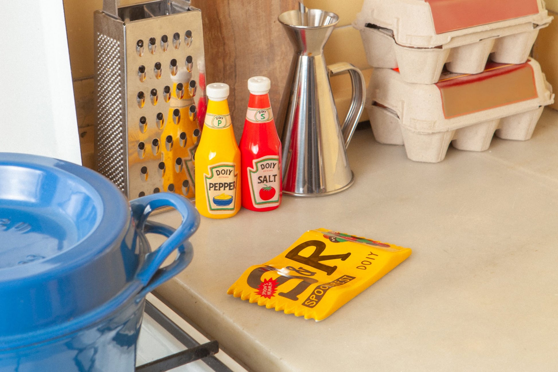 A kitchen counter with a blue pot, vintage box grater, DOIY Corner Shop Ketchup & Mustard Salt & Pepper Set by DOIY, metal oil dispenser, stacked egg cartons, and a cheese wedge yellow sponge adds retro flair.