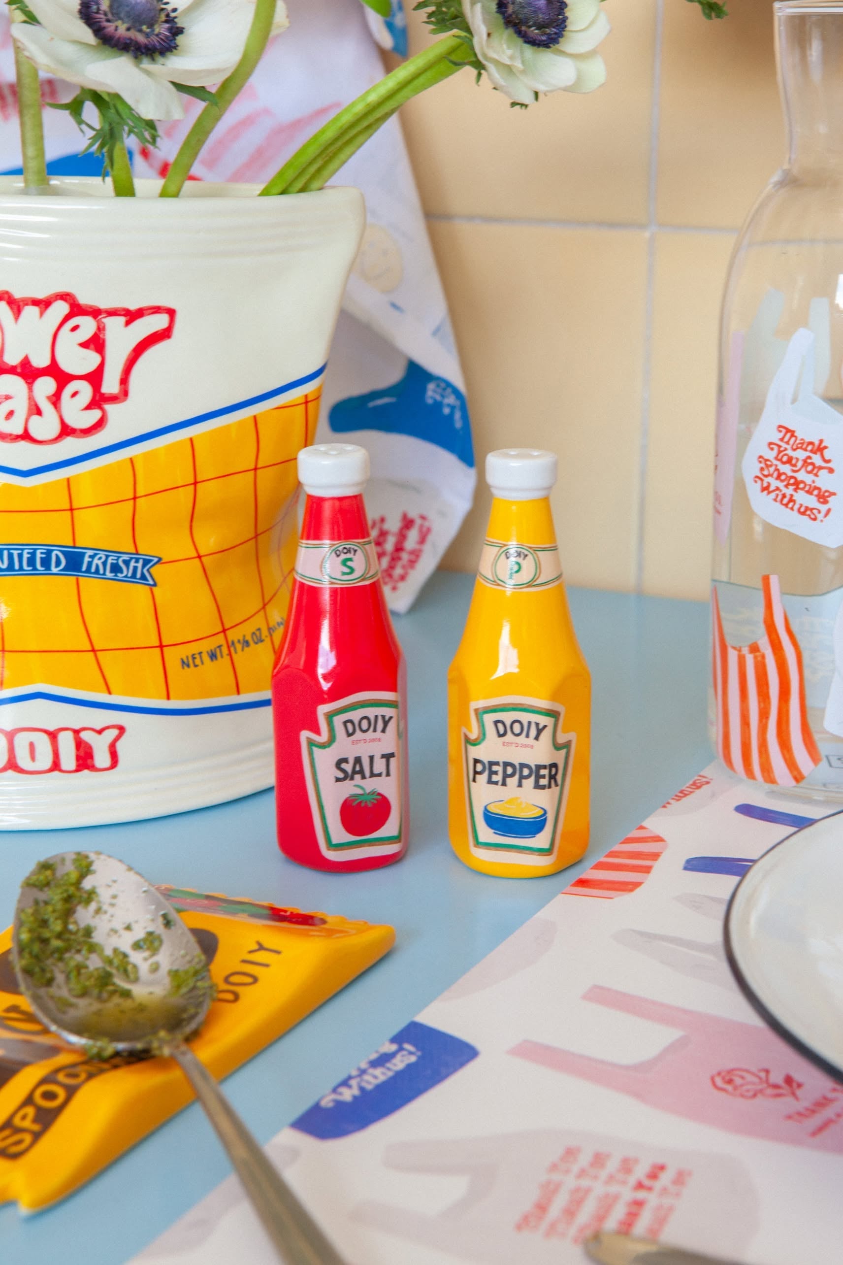 The DOIY Corner Shop Ketchup & Mustard Salt & Pepper Set by DOIY adds retro charm to any colorful countertop, especially when paired with a flower vase, patterned napkin, and glass carafe.