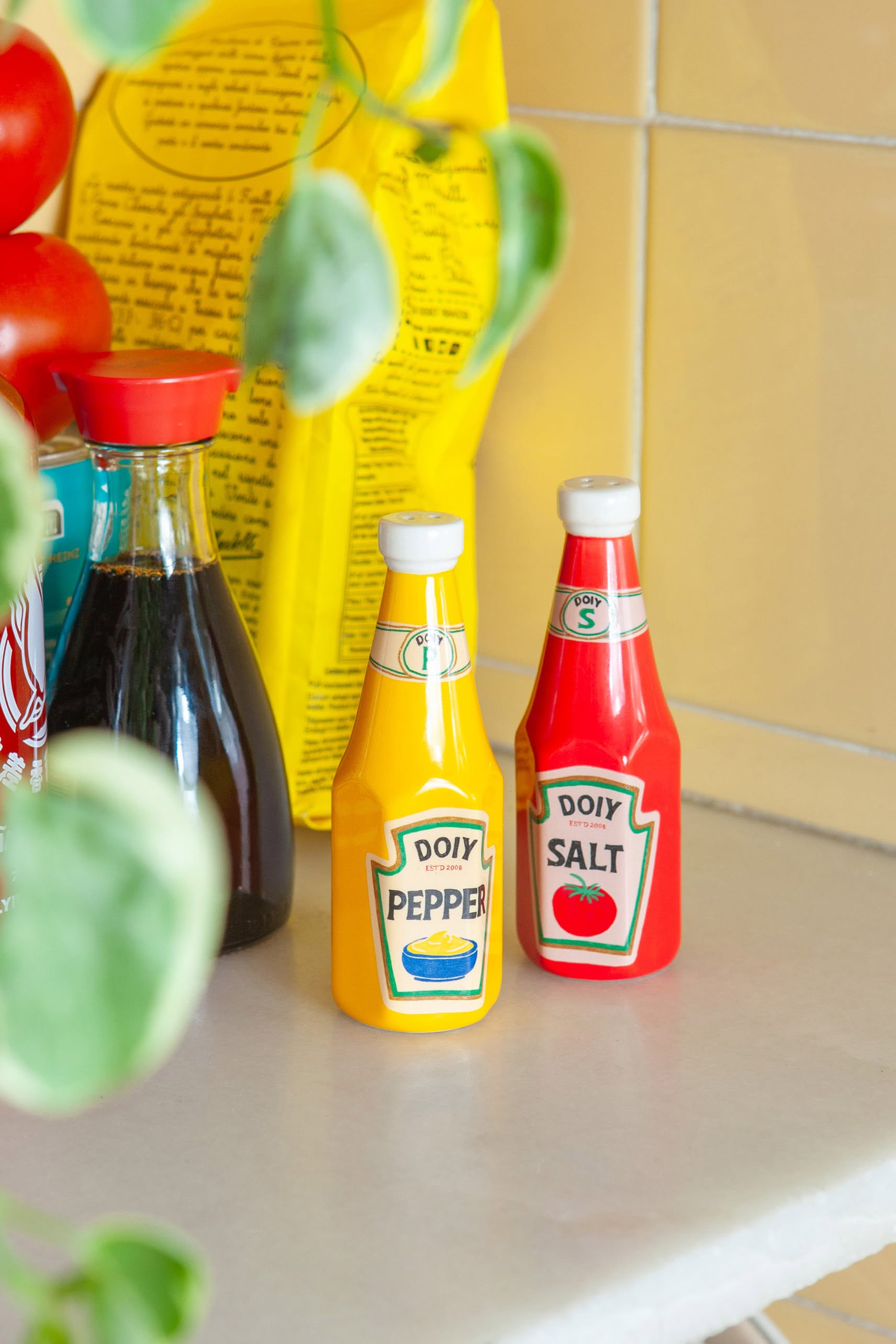 Two DOIY Corner Shop Ketchup & Mustard Salt & Pepper Set bottles bring retro flair to the counter, sitting beside soy sauce, tomatoes, and a yellow package, with a green plant hanging in the foreground.