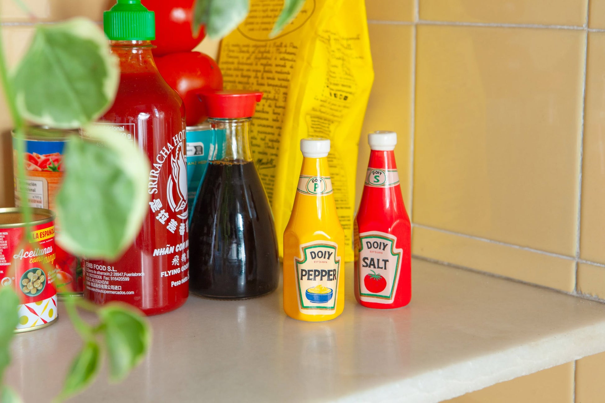 A kitchen counter shows condiments like Sriracha sauce, soy sauce, a can, a yellow pepper shaker, the DOIY Corner Shop Ketchup & Mustard Salt & Pepper Set by DOIY shaped like bottles, and a bag of red tomatoes in the background.
