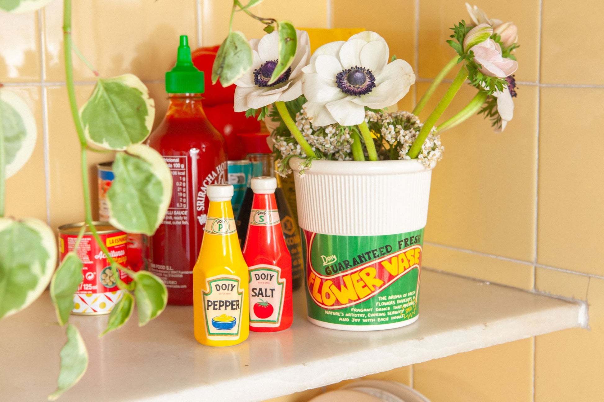 A white cup labeled FLOWER VASE with fresh flowers sits on a retro kitchen counter, surrounded by potted plants, condiment bottles, and the DOIY Corner Shop Ketchup & Mustard Salt & Pepper Set against yellow tile.