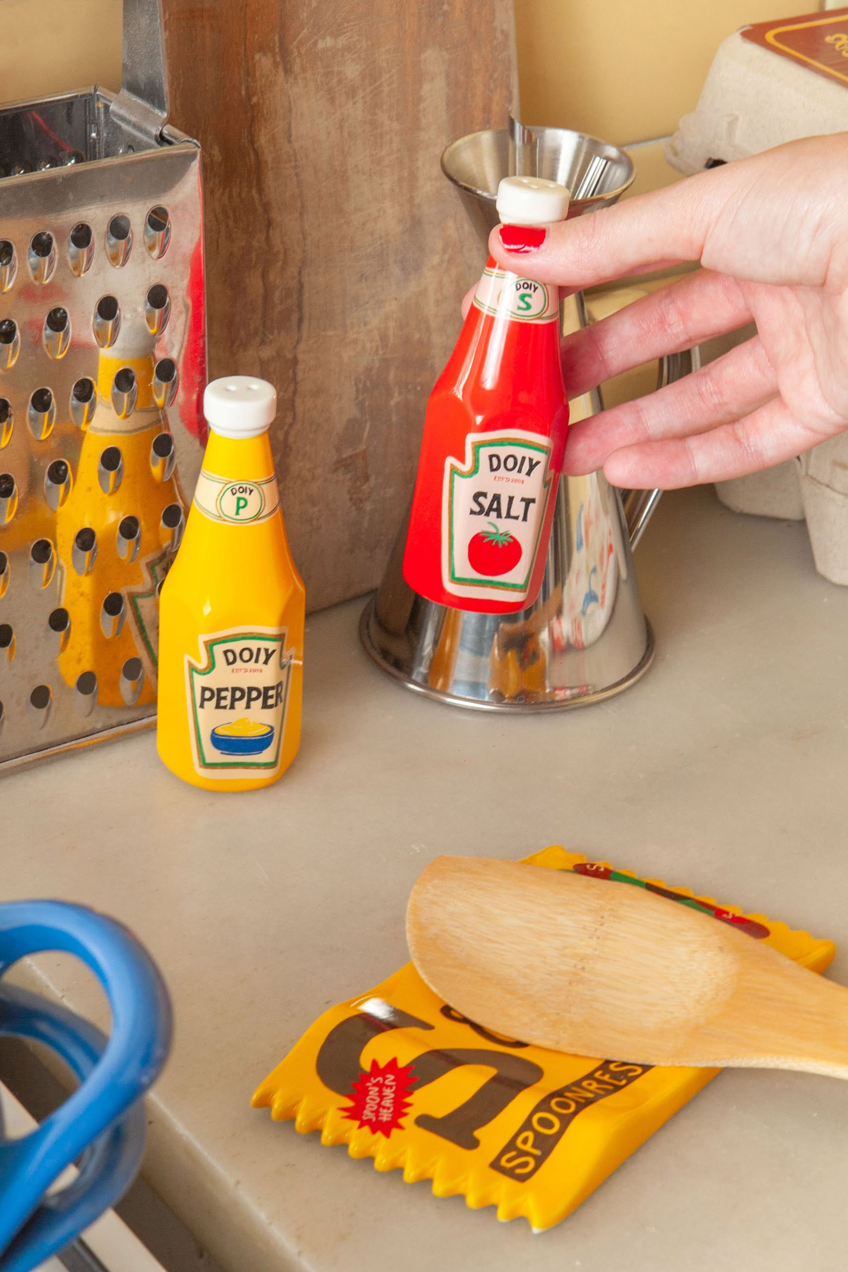 A hand holds the red ketchup-shaped shaker beside the yellow mustard-shaped shaker from the DOIY Corner Shop Ketchup & Mustard Salt & Pepper Set, adding retro flair to a kitchen with a grater, wooden spoon, and metal pitcher.