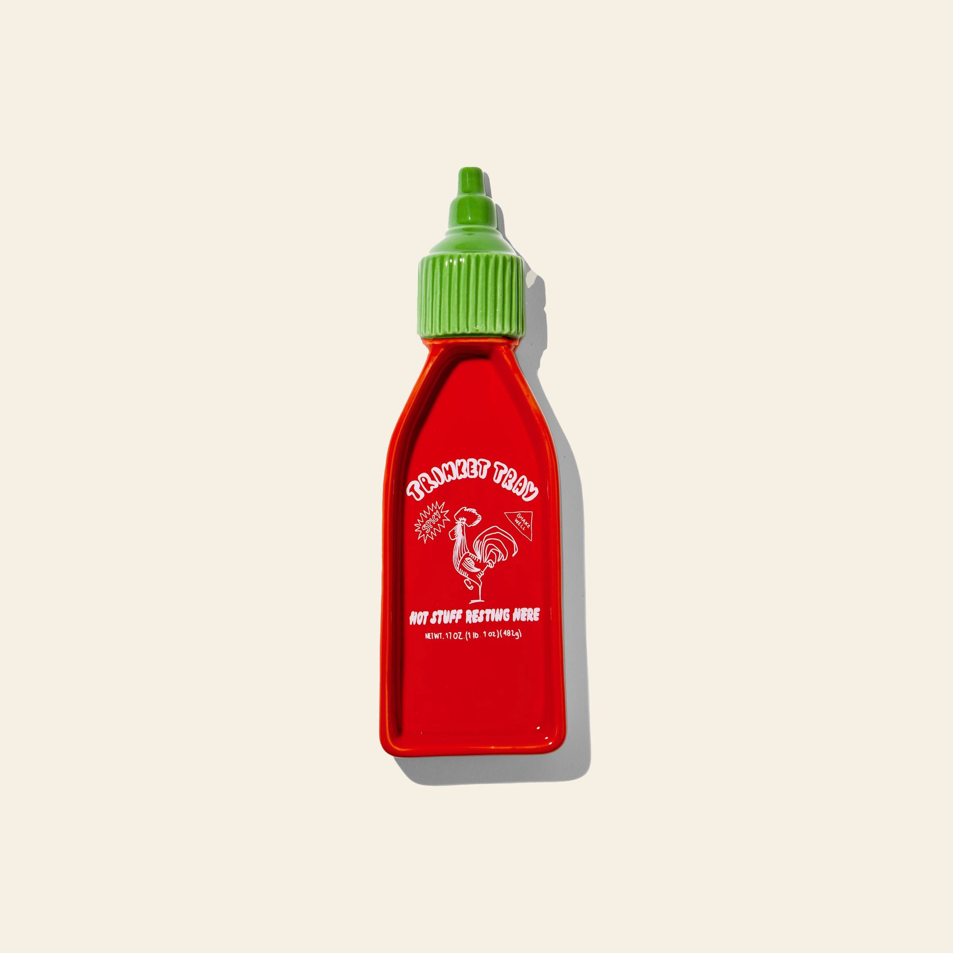 The DOIY Corner Shop Sriracha Trinket Tray by DOIY features a red bottle with a green cap, retro rooster illustration, and nostalgic white text, designed to resemble sriracha sauce. It’s displayed on a light beige background.