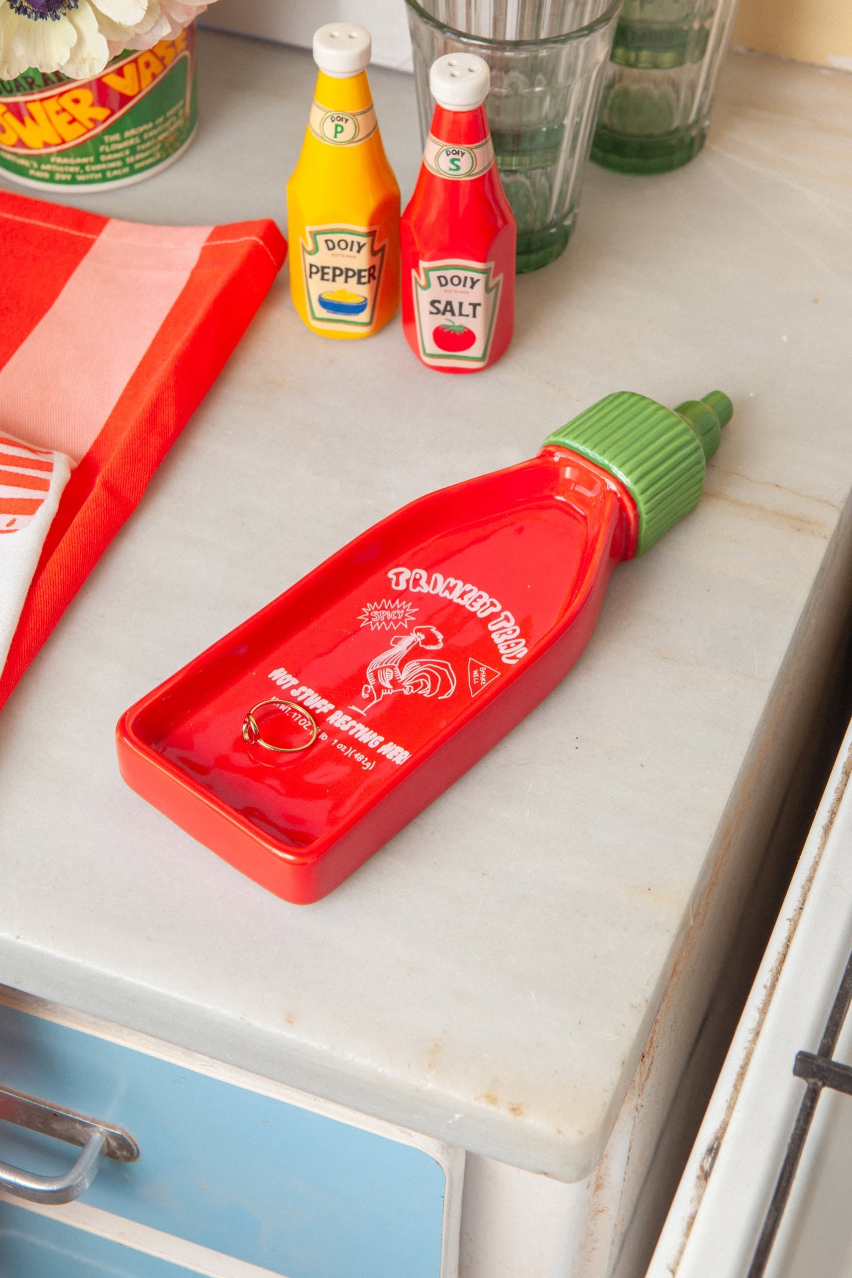The DOIY Corner Shop Sriracha Trinket Tray, a red bottle-shaped piece with nostalgic design by DOIY, sits on a kitchen counter beside a red dish towel and two ceramic condiment containers labeled Doiy Pepper and Doiy Salt.