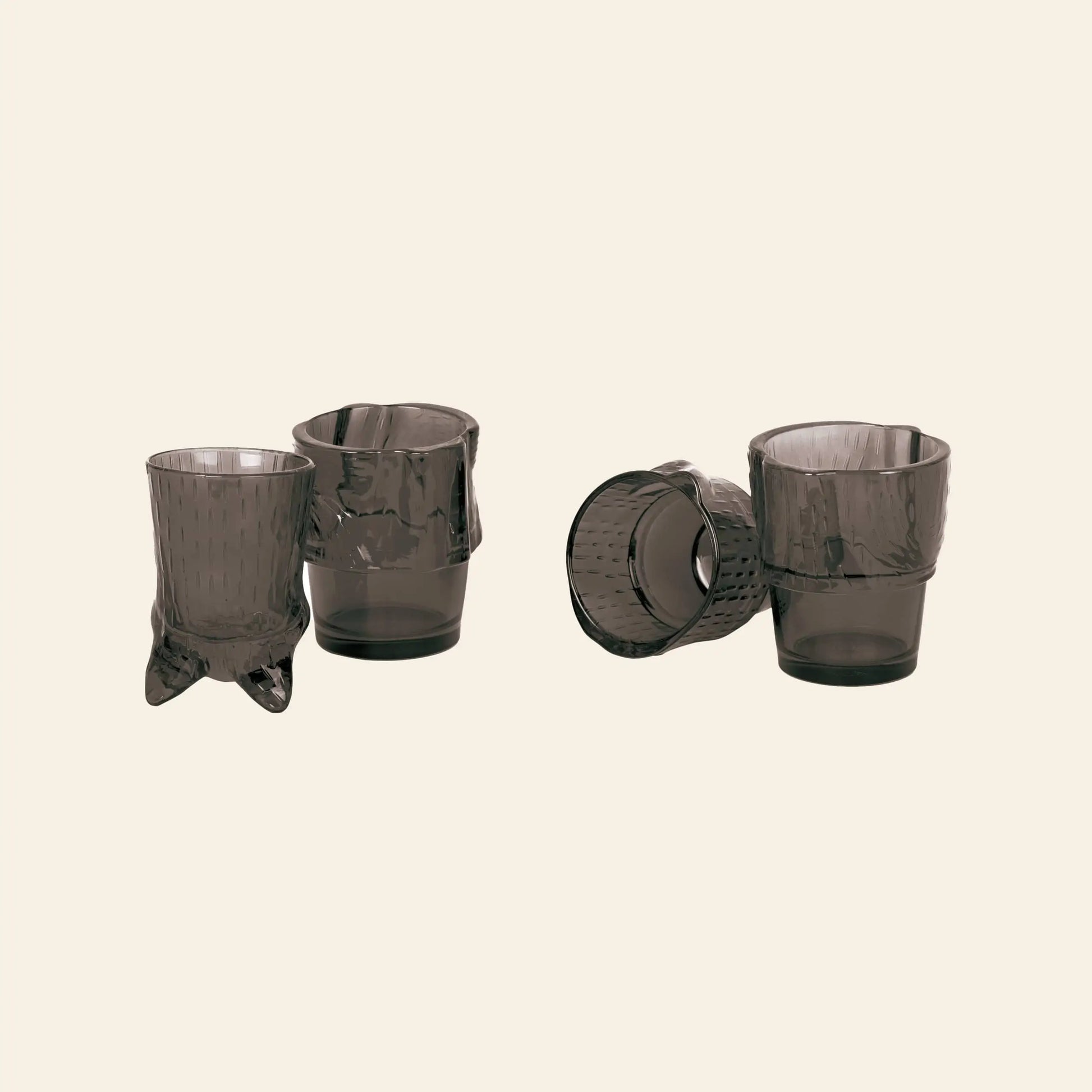 The DOIY Kitty Glasses set includes four smoky gray glasses with unique, crumpled shapes. Arranged in pairs on a beige background, two stand upright and two lie on their sides, creating eye-catching decorative pieces.