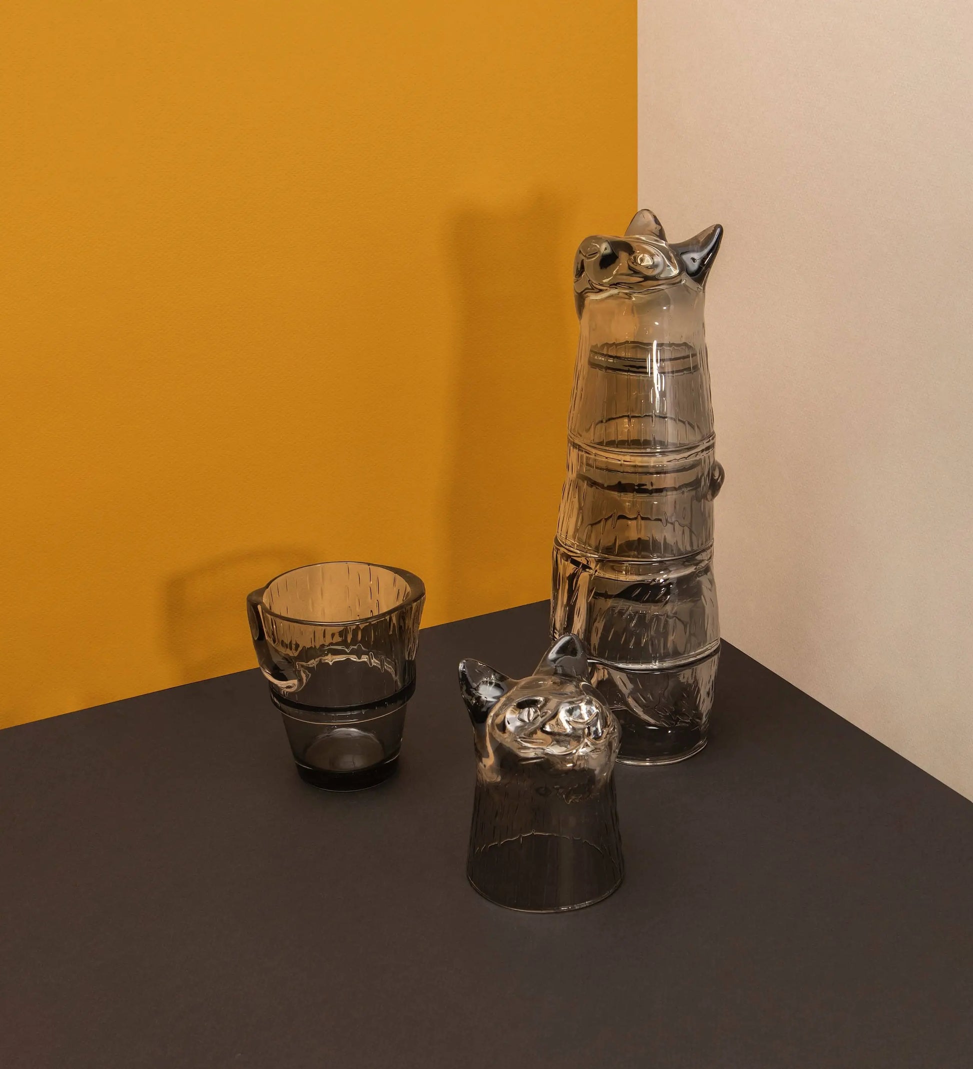 Four stackable DOIY Kitty Glasses, each shaped like a cat face, are arranged on a dark surface. Three pieces form one tall cup. The diagonally split background features mustard yellow and light beige. Brand: DOIY.