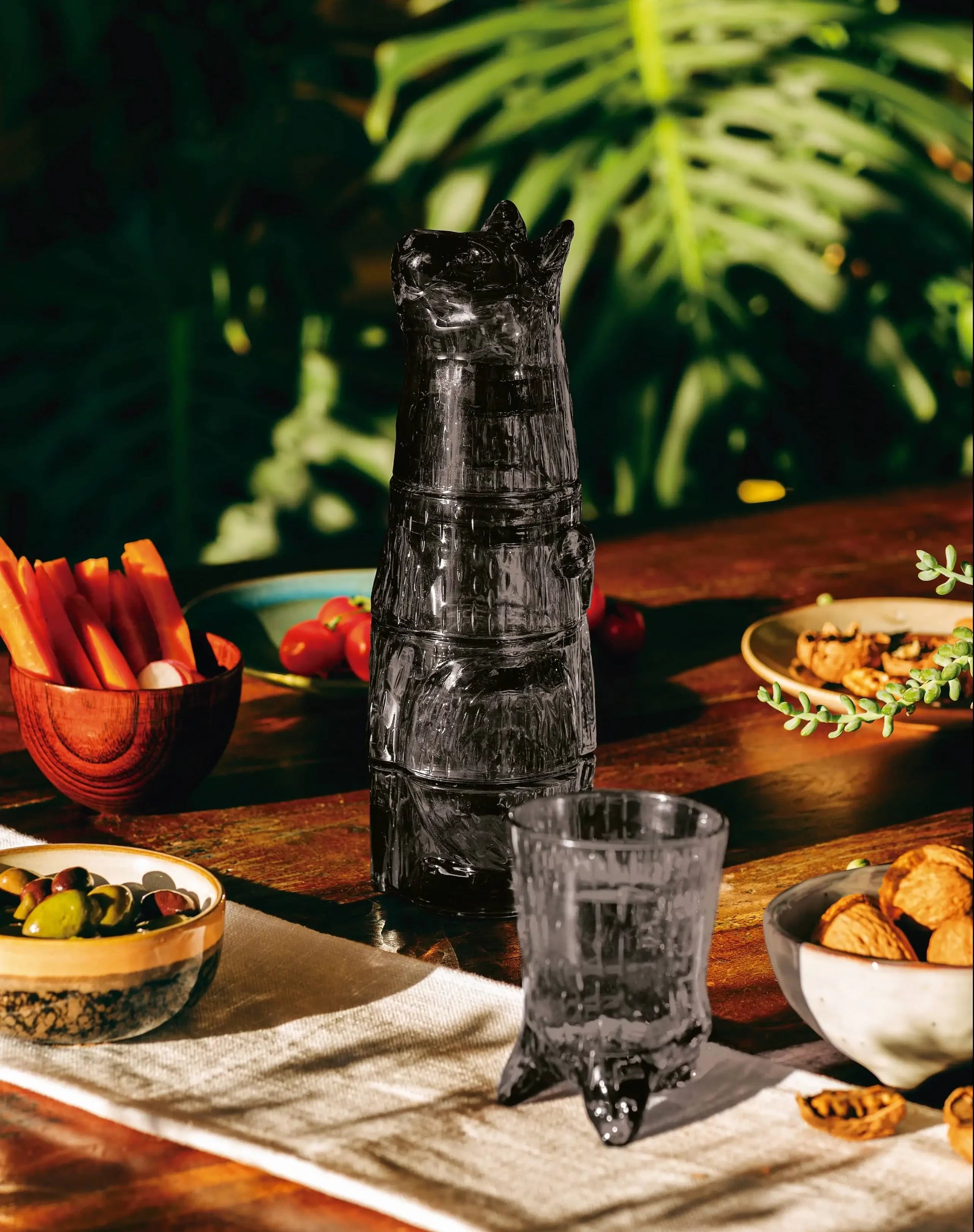 A dark animal-shaped glass carafe sits on a wooden table beside a DOIY Kitty Glasses, Set of 4. The table features bowls of snacks and vegetables, with green leaves in the background.