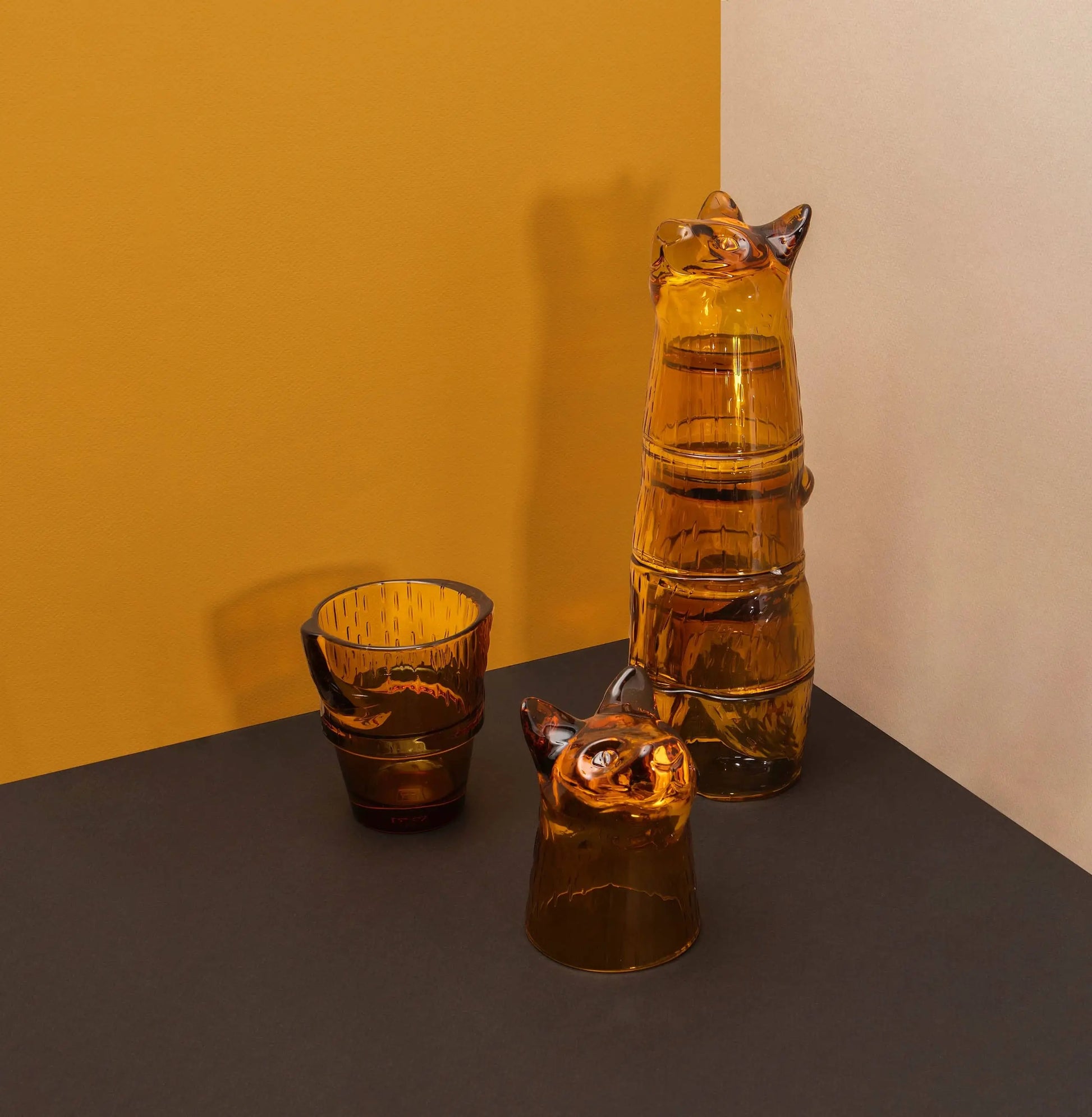 A set of four DOIY Kitty Glasses by DOIY, made from amber glass and stackable, are displayed on a dark surface with yellow and beige walls—charming decorative pieces for any space.