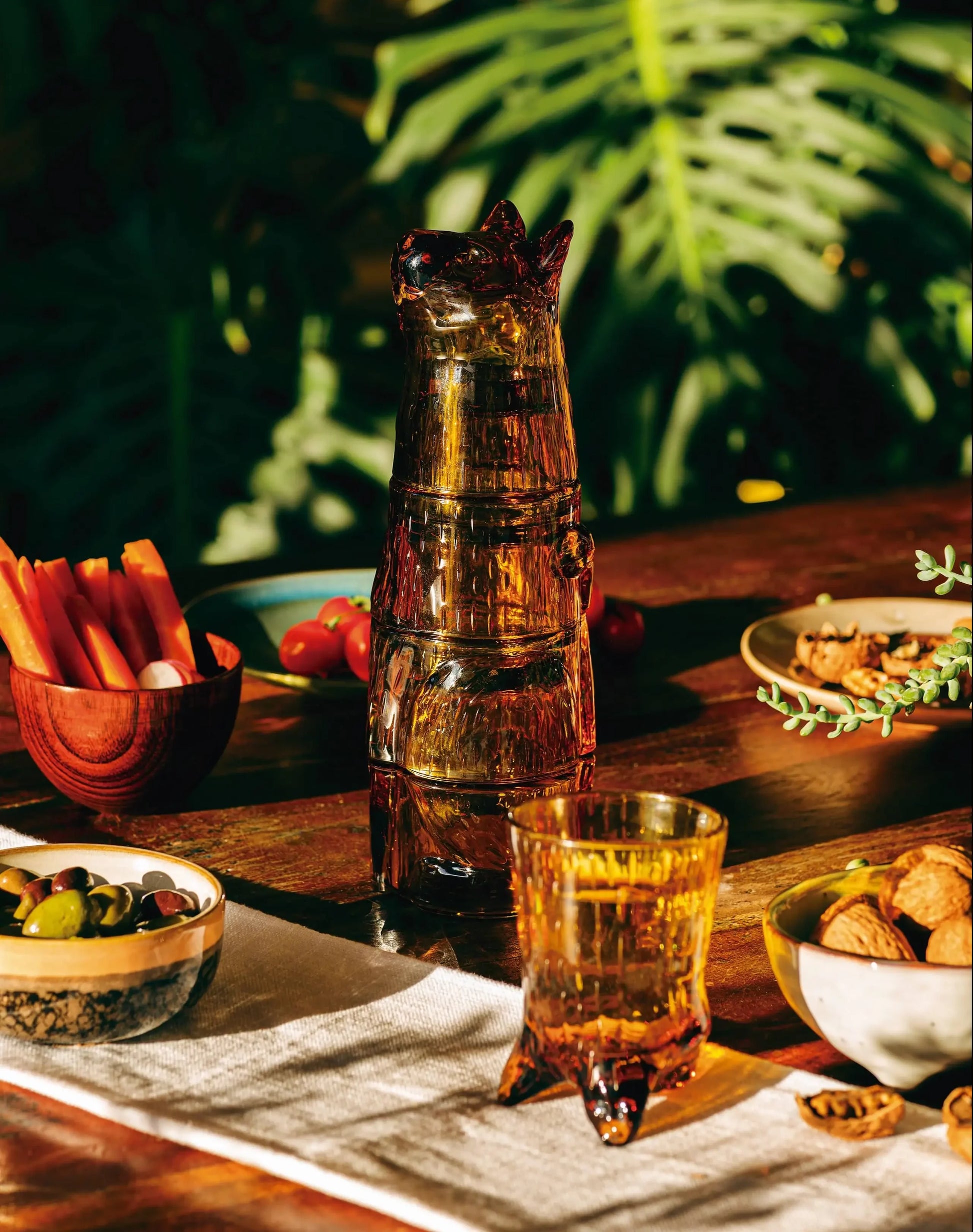 The DOIY Kitty Glasses, Set of 4 by DOIY are stacked on a wooden table with bowls of vegetables, olives, and nuts, while green leafy plants brighten the sunlit background.