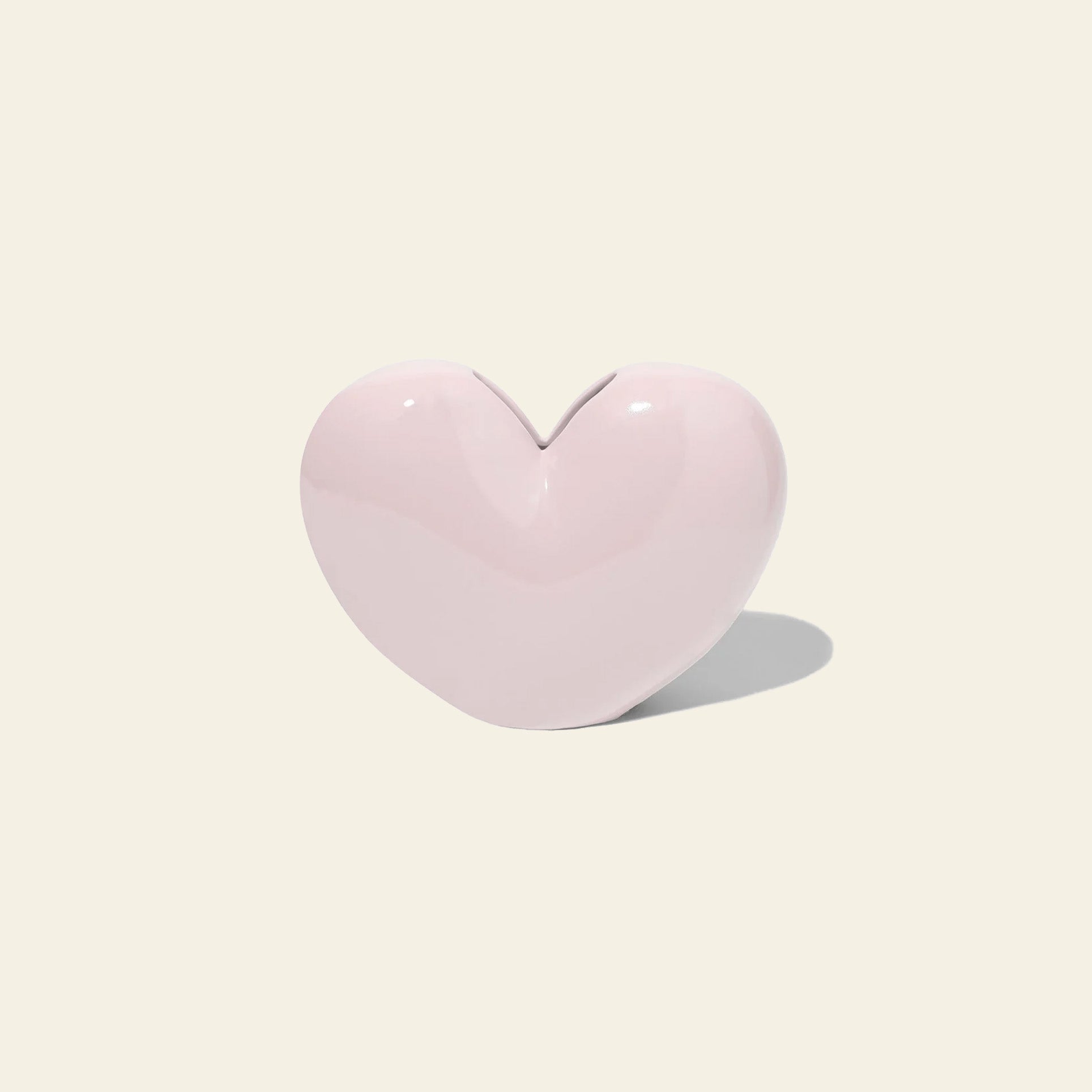 The DOIY Love Vase by DOIY, a glossy light pink heart-shaped ceramic piece, sits on an off-white background casting a soft shadow—an elegant symbol of timeless beauty.