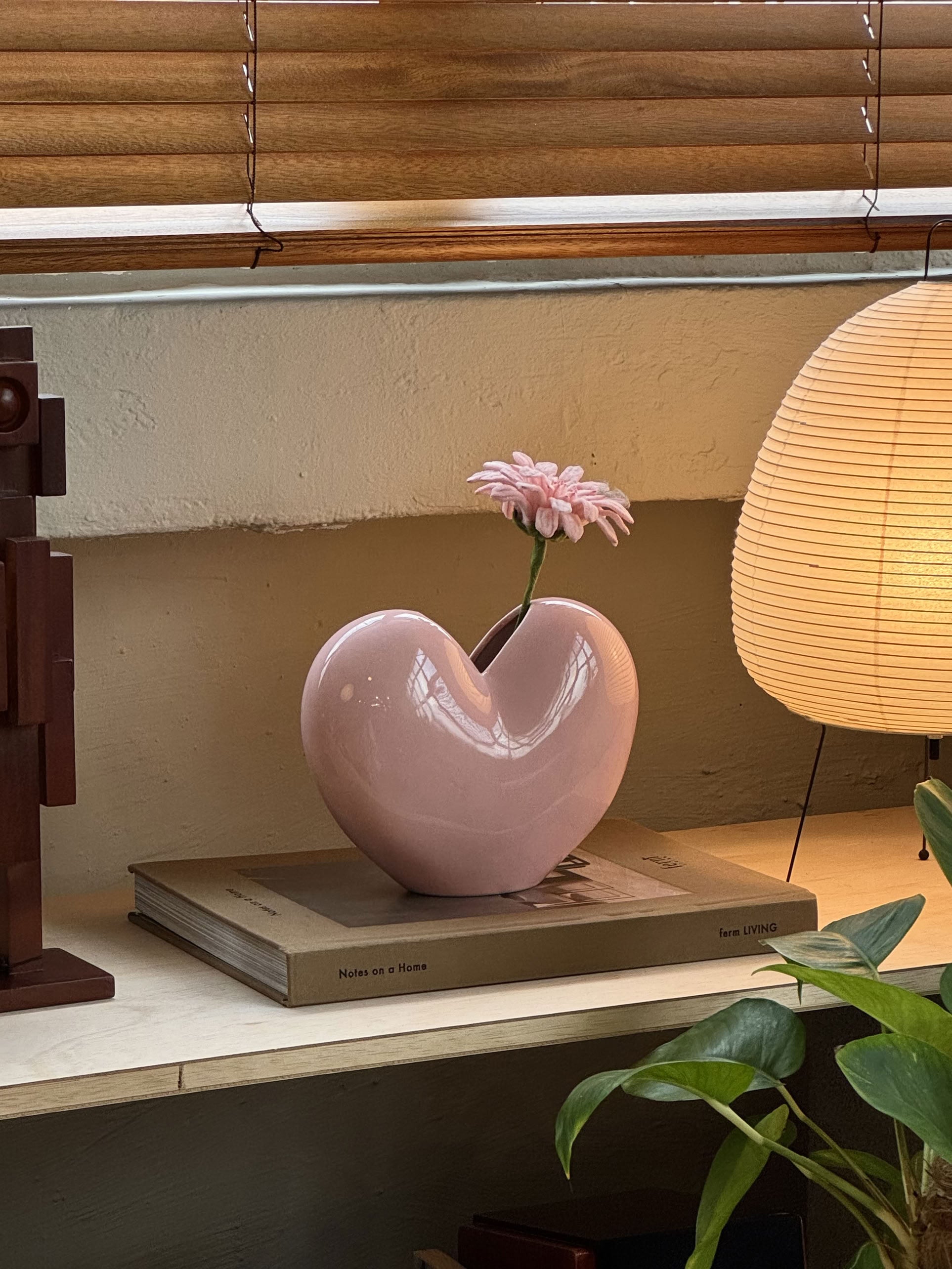 The DOIY Love Vase, a heart-shaped pink accent by DOIY, with a single pink flower, rests on a book beside a round lamp and green potted plant, its charming design glowing softly beneath wooden window blinds.