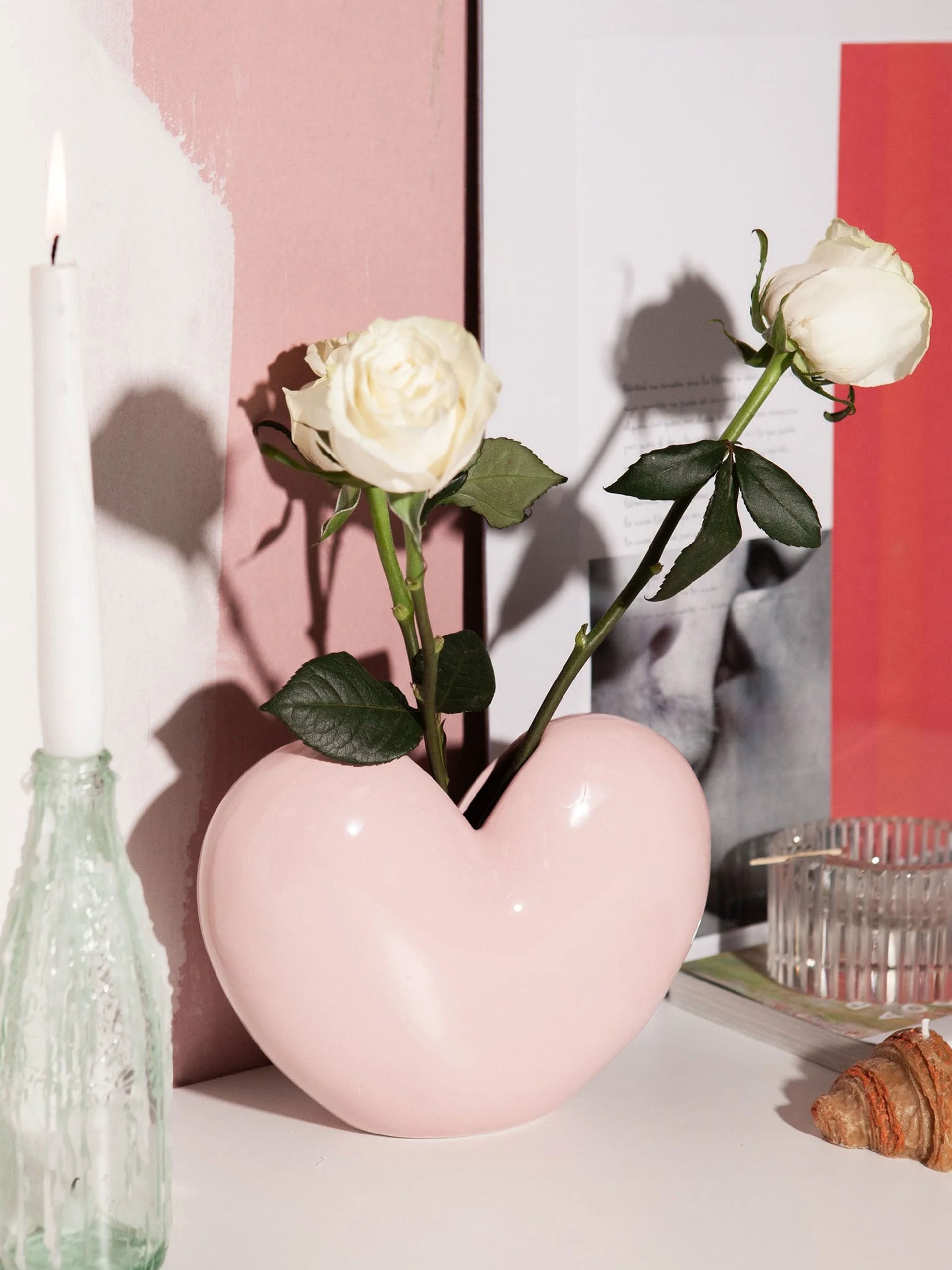 A pink DOIY Love Vase by DOIY holds two white roses on a table with a croissant, lit candle in a textured holder, and framed artwork in the background.