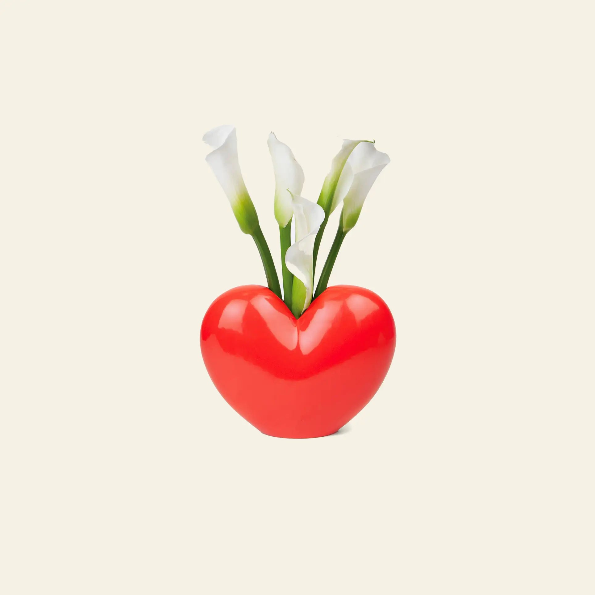 The DOIY Love Vase by DOIY, in red and heart-shaped design, displays several white calla lilies with green stems, highlighting their elegance against a simple light beige backdrop.