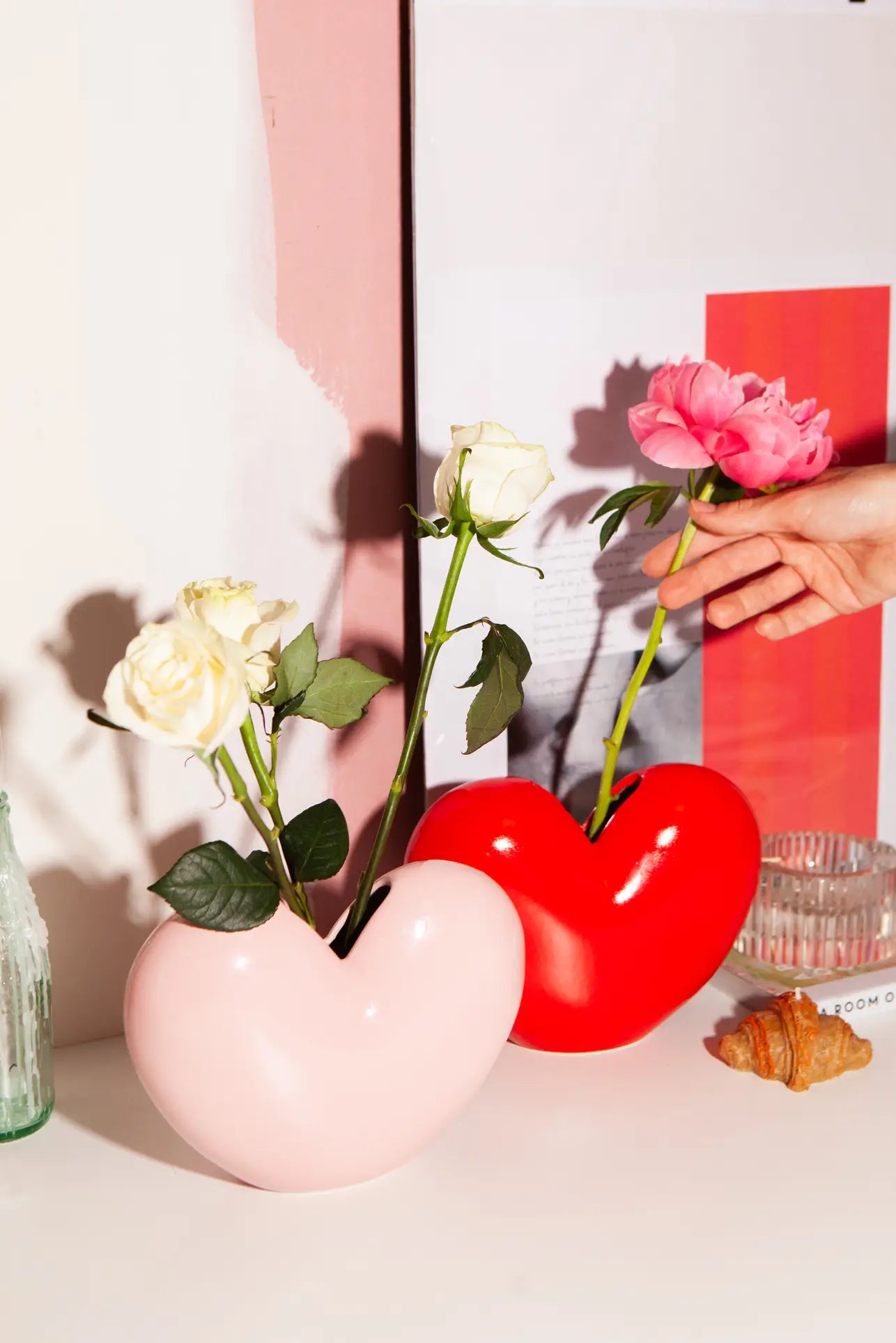 Two DOIY Love Vases—one pink with white roses, the other red with a pink peony—by DOIY sit on a white surface, surrounded by glass decor and a seashell, showcasing sentimental design and timeless beauty.