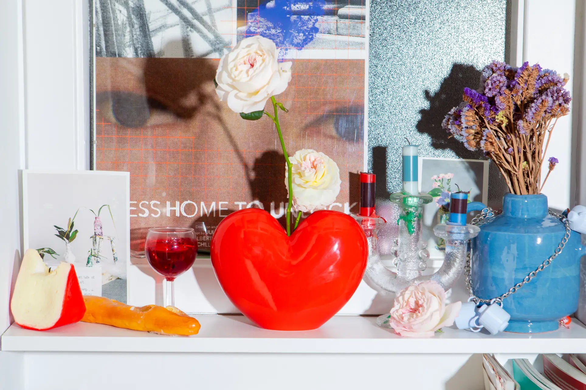 A colorful shelf display features the DOIY Love Vase—a red, heart-shaped vase by DOIY with white roses—next to a wine glass with red liquid, cheese, a blue vase of dried flowers, candles, a postcard, and decor against an art print backdrop.