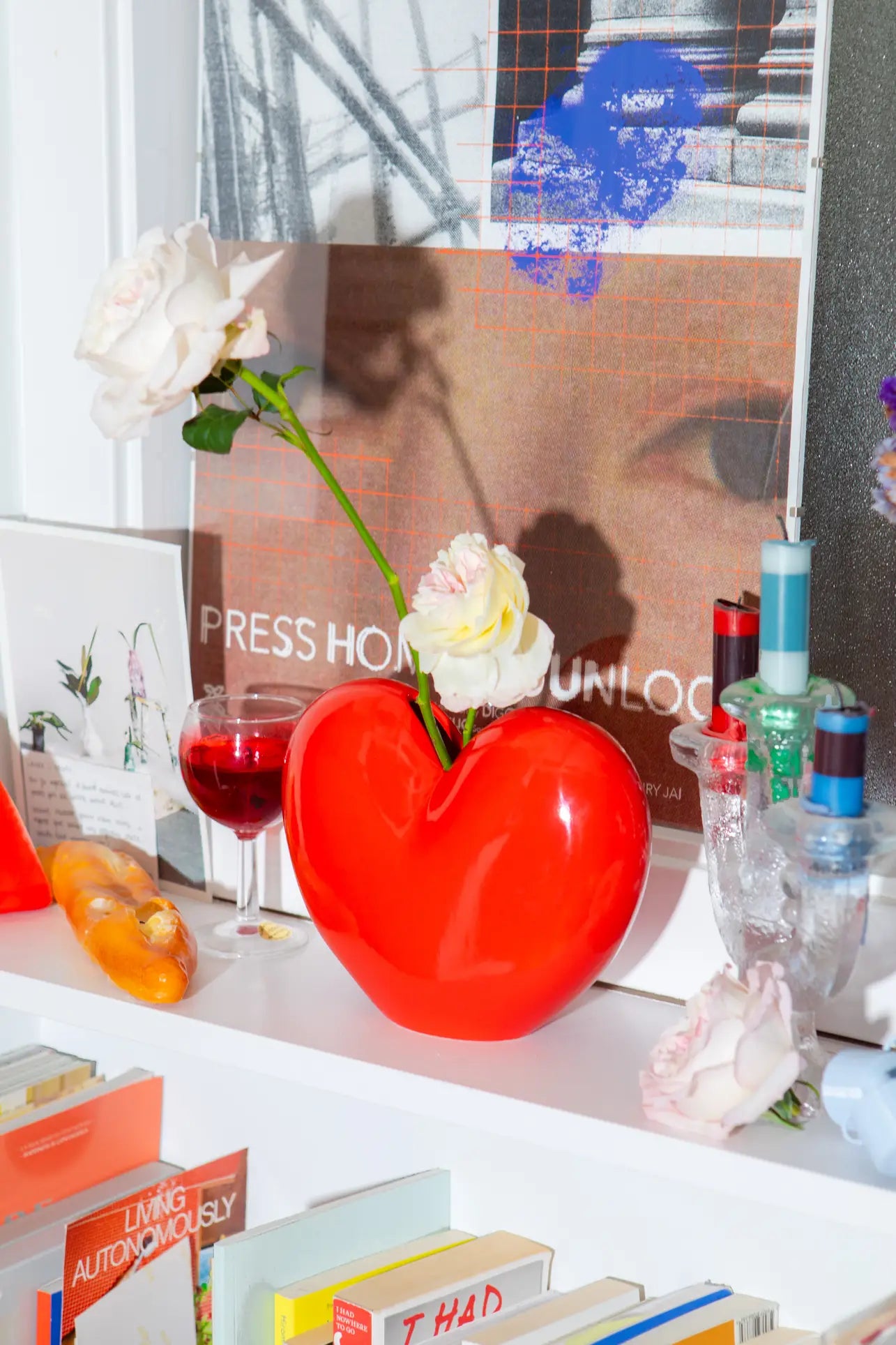 The DOIY Love Vase by DOIY, in vibrant red and filled with white roses, stands on a shelf among books, red wine, trinkets, and art prints—creating a lively display that honors sentimental design and timeless beauty.