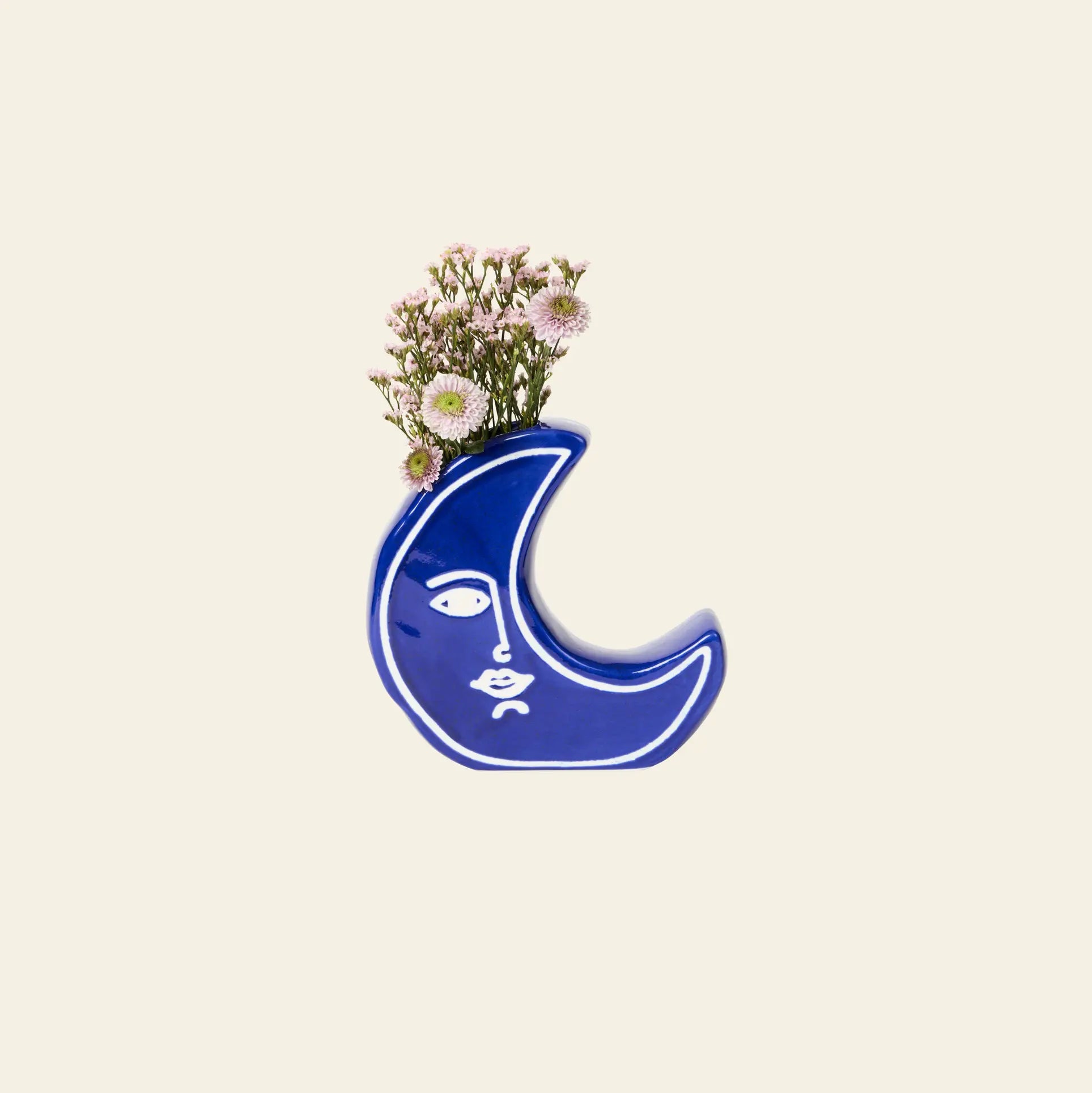 The DOIY Moon Vase by DOIY features a blue finish with a white abstract face and displays a small bouquet of light pink flowers, adding celestial charm to a simple beige backdrop.