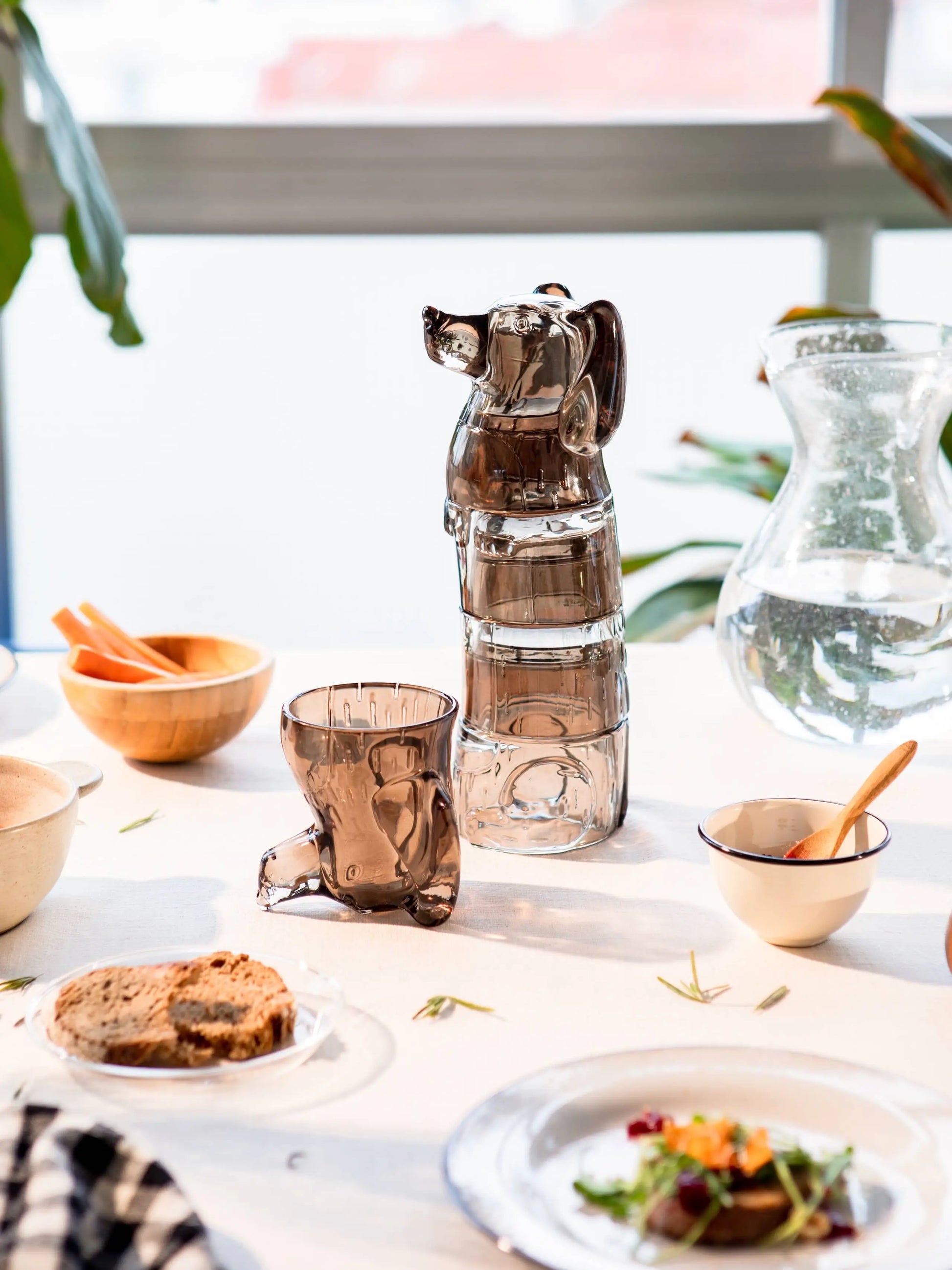 A set of DOIY Puppy Glasses (set of 4) by DOIY sits among decorative items, bowls, stackable plates of food, a glass water pitcher, and leafy plants. Sunlight streams through a large window in the background.