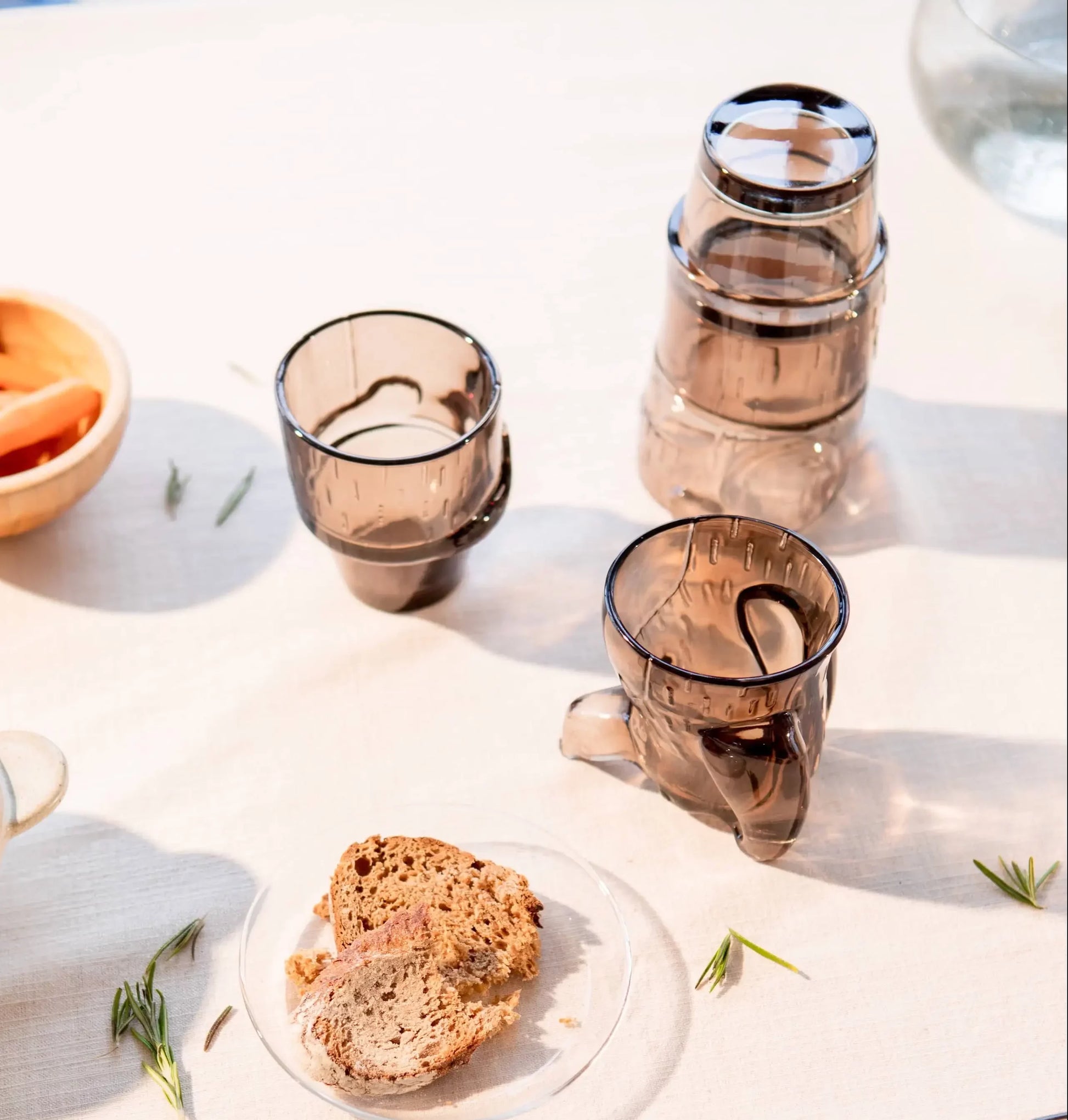 A table set with DOIY Puppy Glasses (set of 4, translucent brown) by DOIY, a clear plate of bread slices, a bowl of orange slices, and rosemary sprigs on a light tablecloth in soft sunlight.