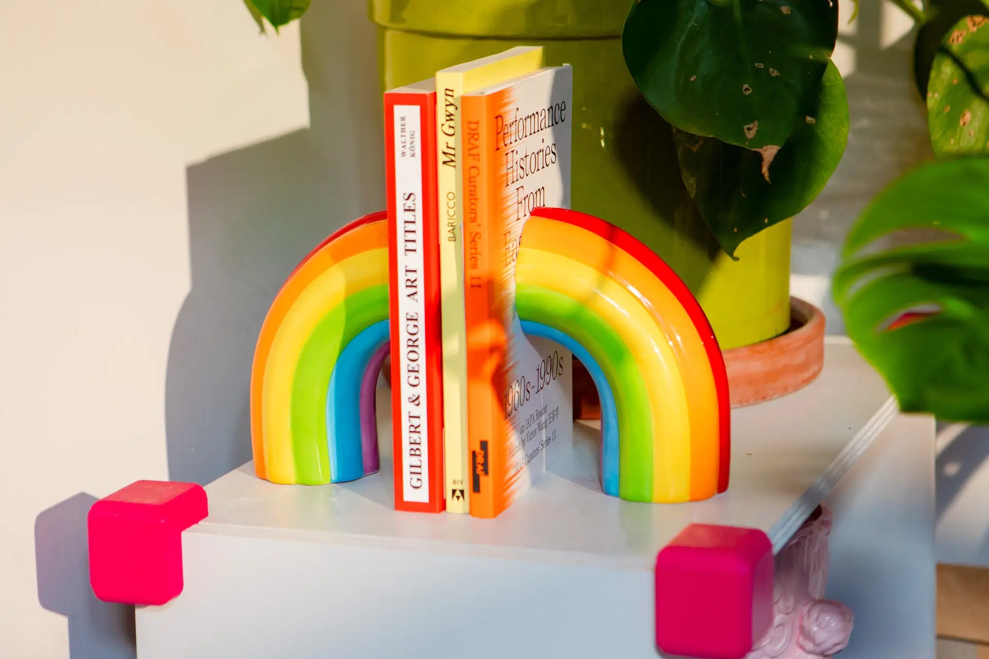 The DOIY Rainbow Bookend by DOIY brings a splash of color to your decor, holding up to three books on a white table surrounded by green plants as sunlight creates warm shadows.