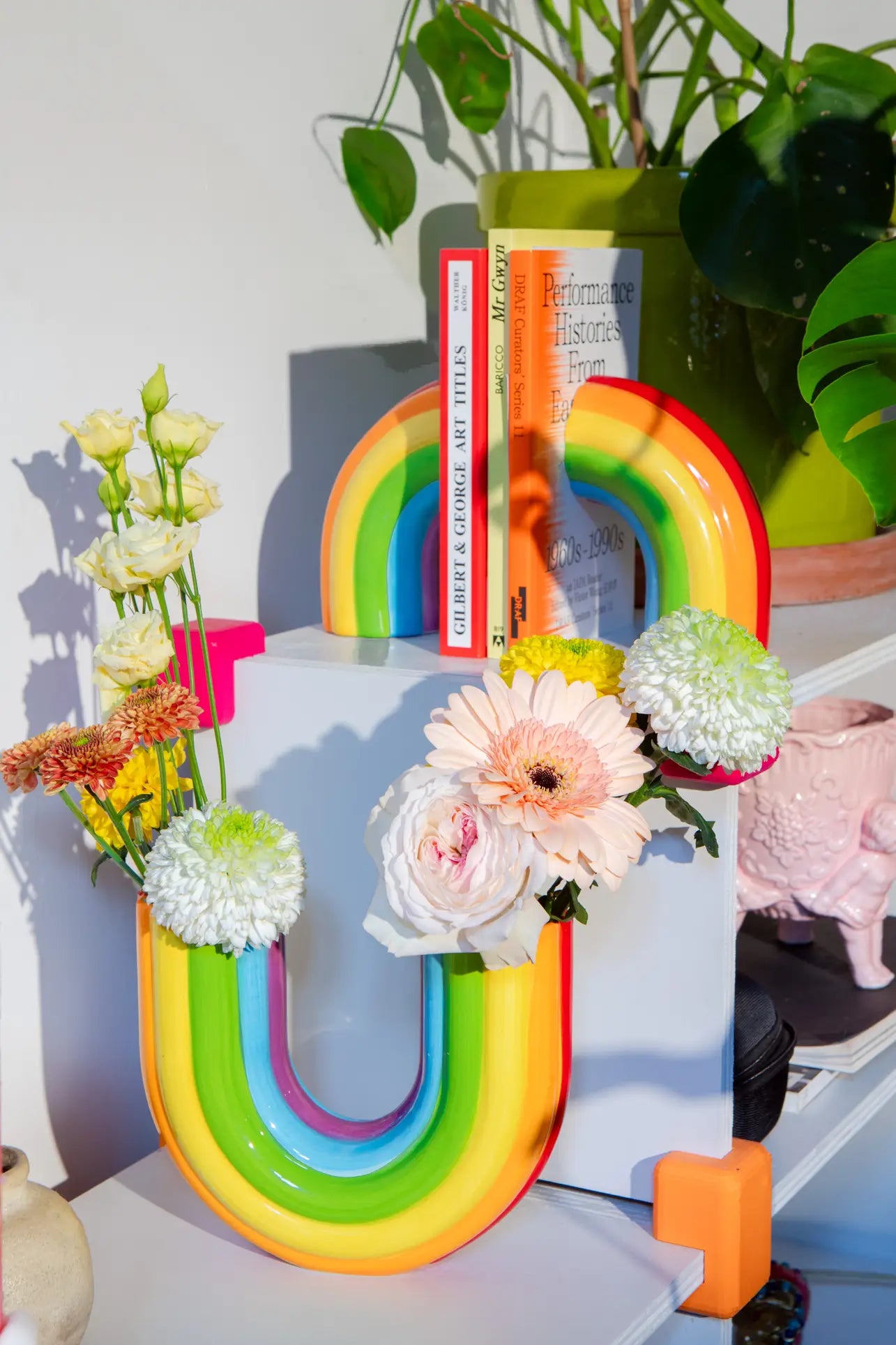 Colorful U-shaped vases with rainbow stripes display assorted flowers, while DOIY Rainbow Bookends by DOIY and green plants decorate a white and gray shelf, all bathed in sunlight for a cheerful home decor look.