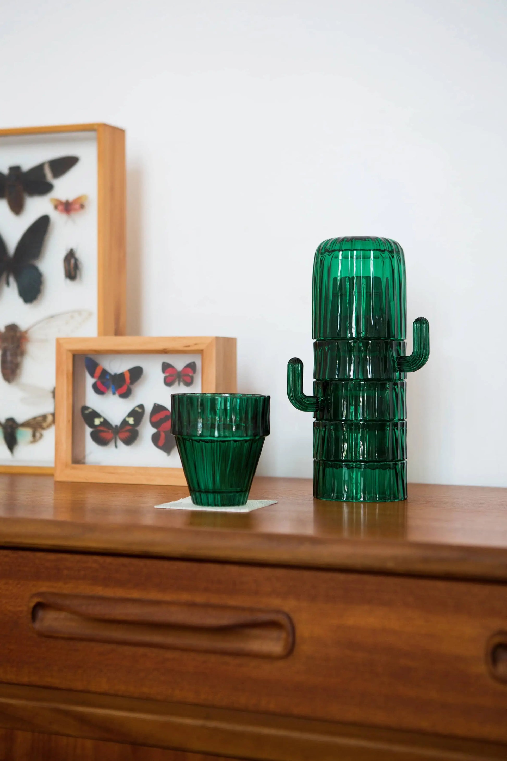 DOIY Saguaro Glasses, Set of 6 by DOIY, are arranged on a wooden dresser. Framed butterfly and moth specimens in the background complete this distinctive home decor scene.