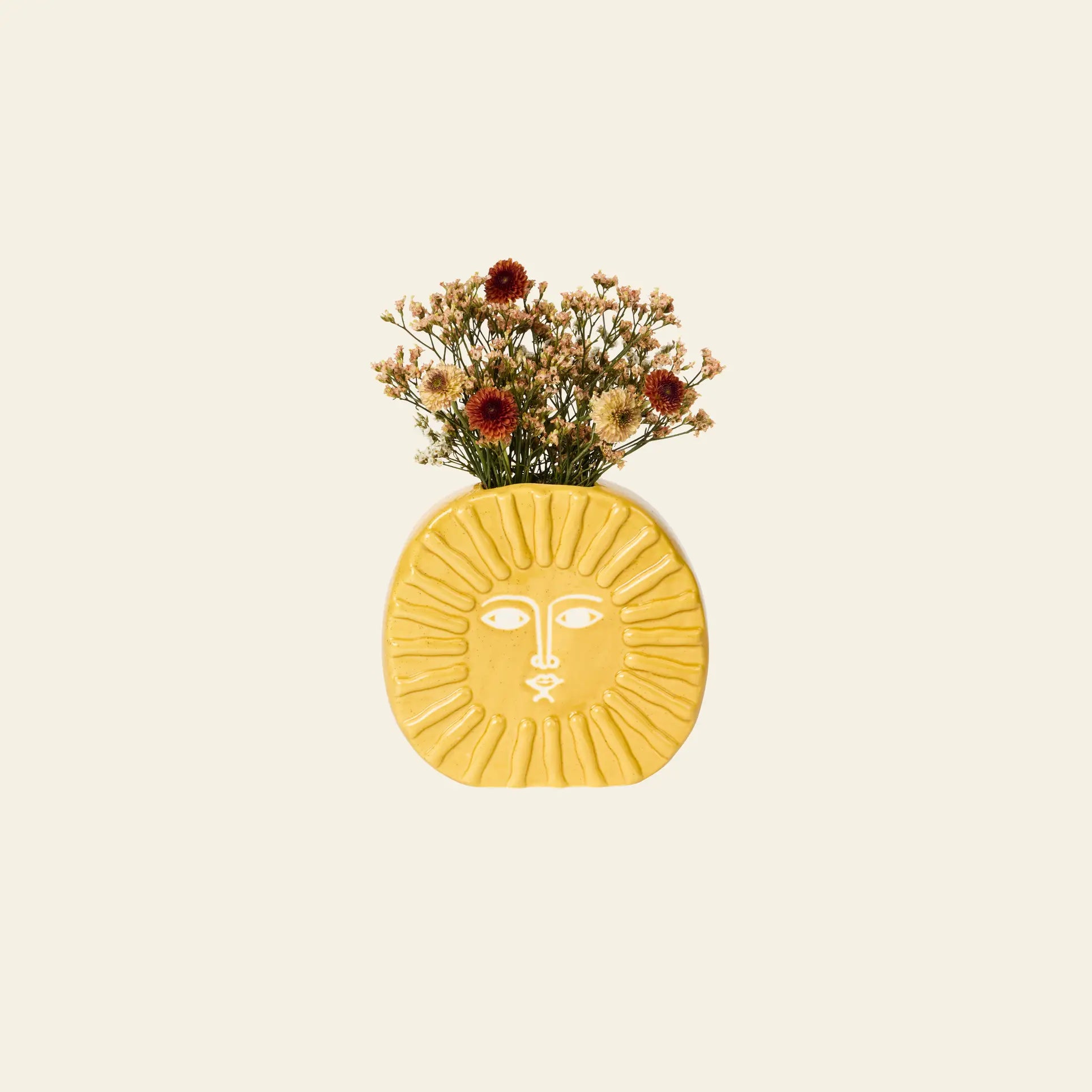 The DOIY Sun Vase by DOIY, a yellow face-shaped vase, adds brightness to your decor and holds dried white and dark red flowers against a simple light background.