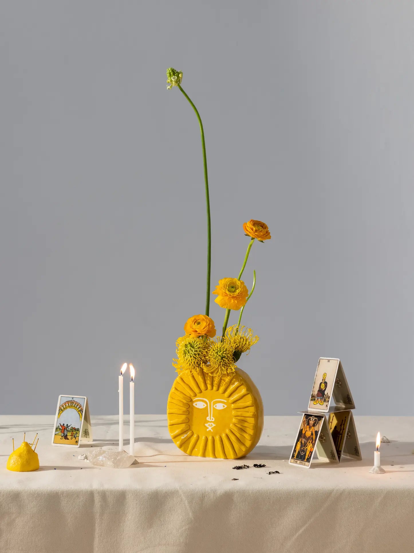 The DOIY Sun Vase by DOIY, a yellow ceramic vase with a sun-shaped face, holds orange and yellow flowers on a table with tarot cards, candles, a lemon pincushion, and crystals—eye-catching decor on a plain gray background.
