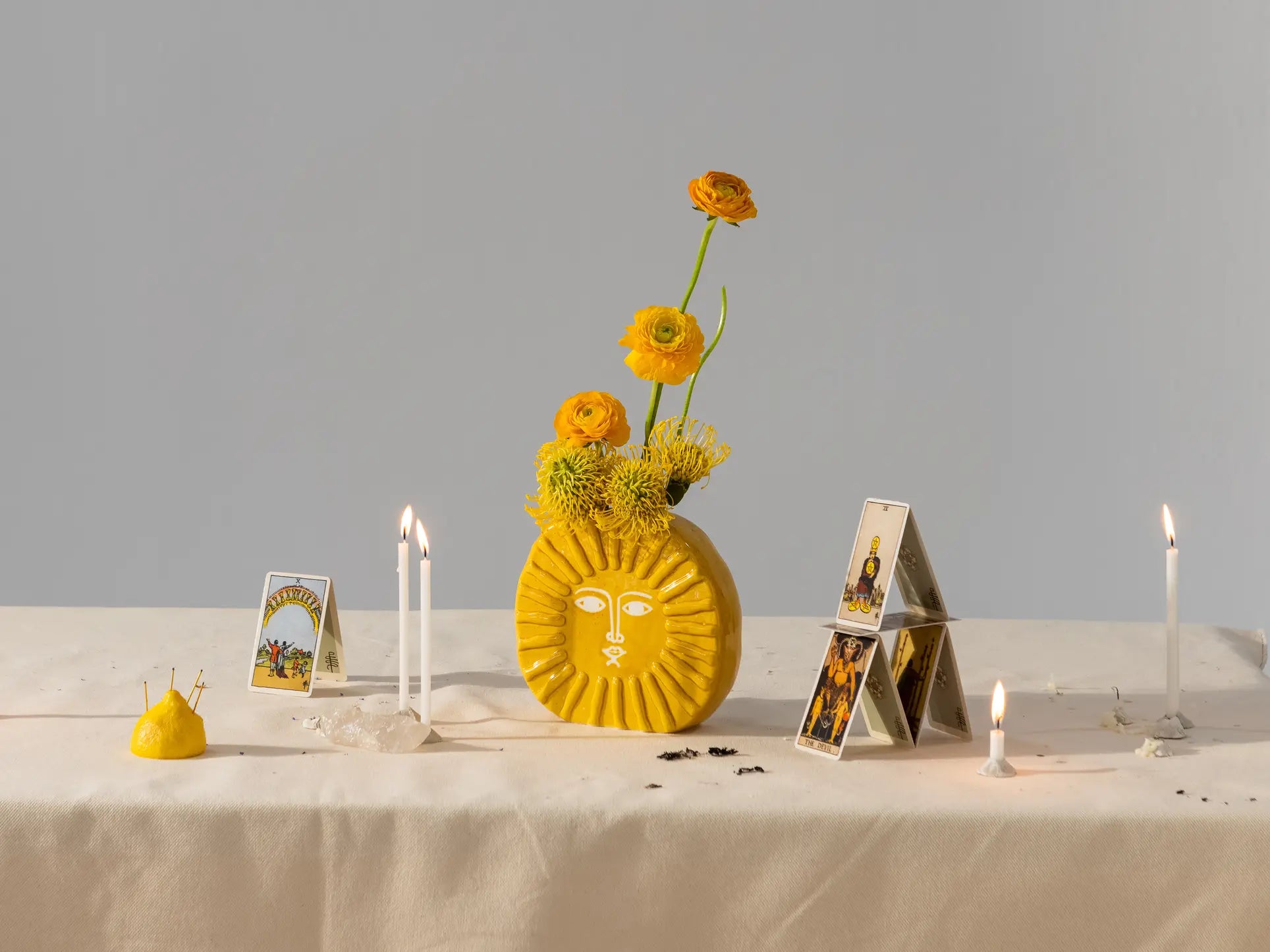 The DOIY Sun Vase by DOIY, filled with flowers, is styled on a cream cloth with tarot cards, lit white candles, crystals, and a yellow pincushion—chic home decor against a plain gray background.