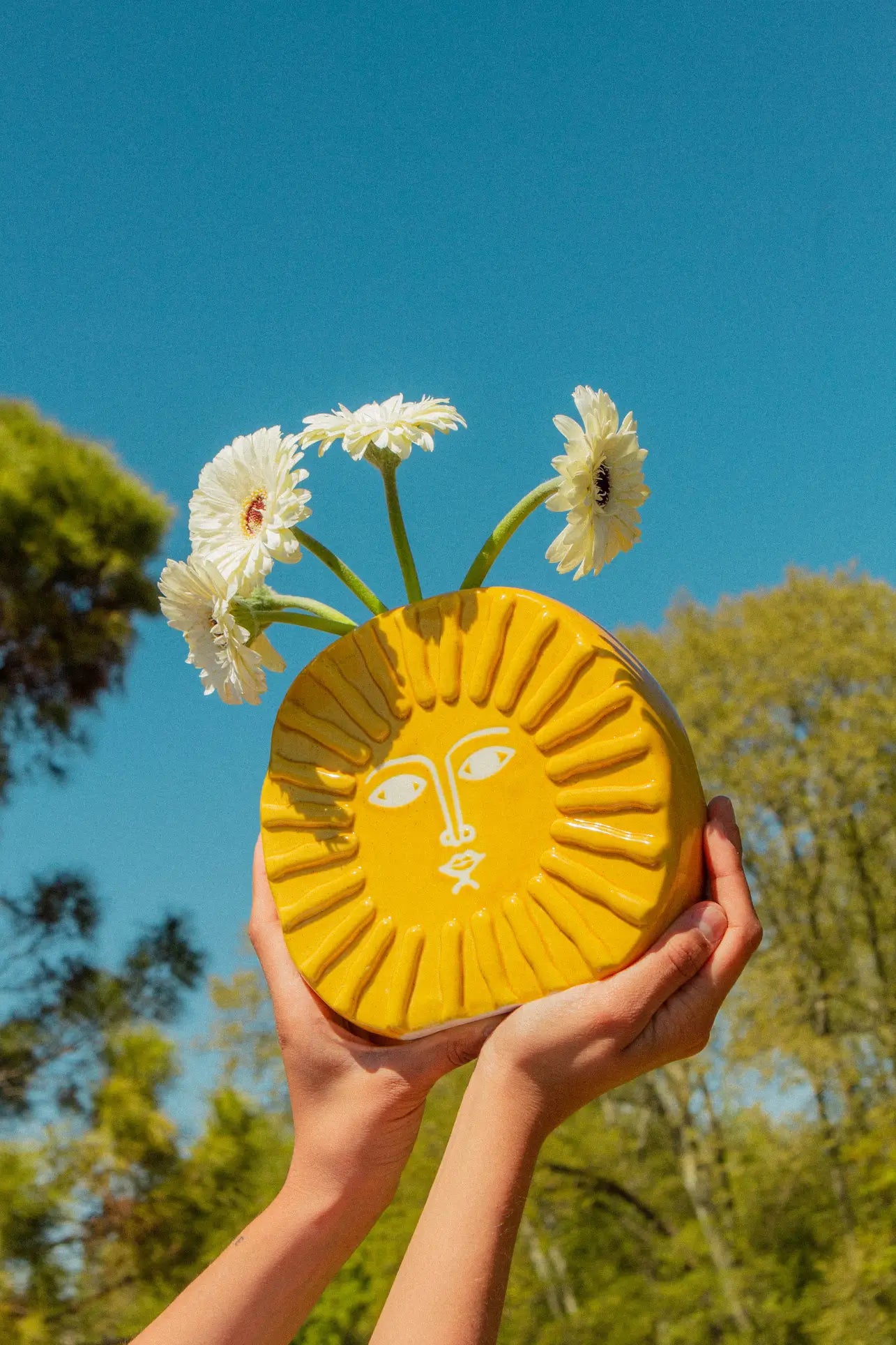 Hands hold up the DOIY Sun Vase by DOIY, featuring a face design in yellow and filled with three white flowers, set against a clear blue sky and green trees—a charming home decor accent.